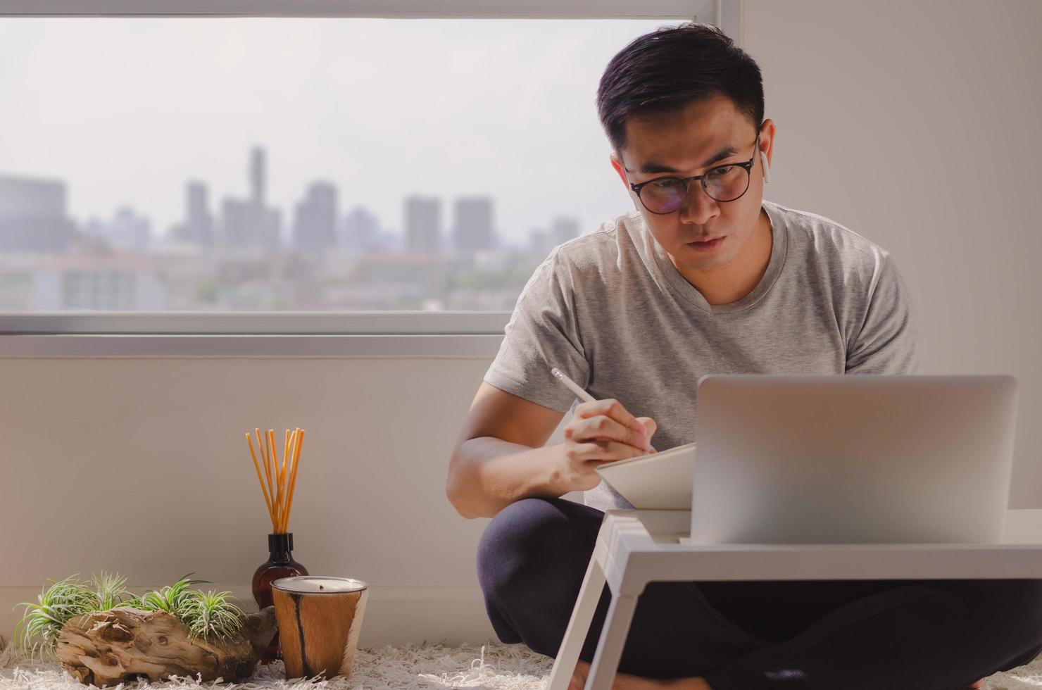 uomo asiatico in abbigliamento casual seduto e al lavoro su un laptop in camera che ha candela, diffusore di aromi e impianto di tillandsia sul pavimento. concentrato sul lavoro quando si lavora a casa. foto