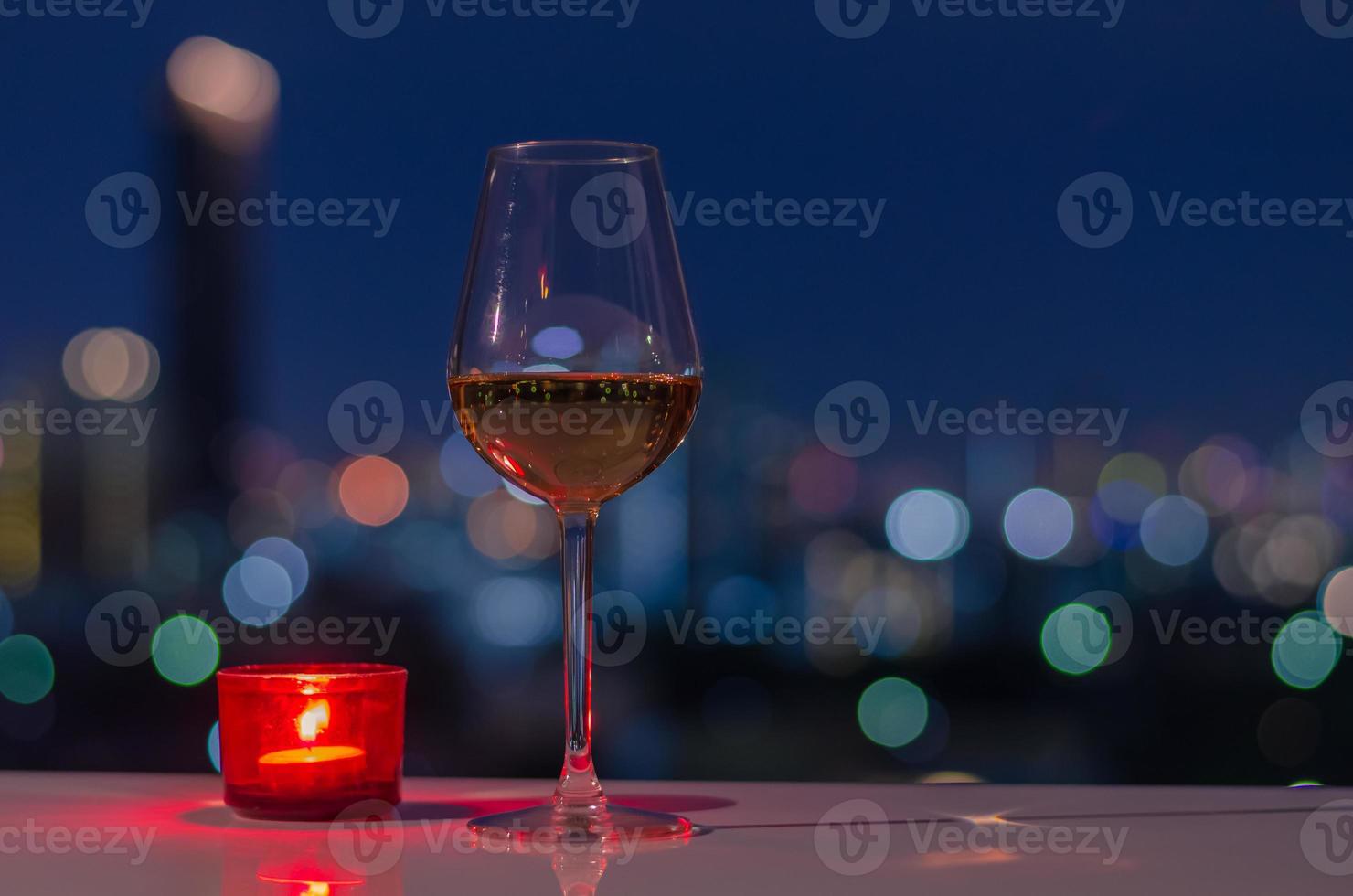 un bicchiere di vino rosato con una candela rossa sul tavolo e sullo sfondo colorato della luce del bokeh della città. foto