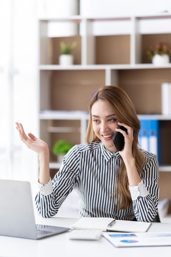 giovane donna d'affari al telefono in ufficio. donna d'affari che manda un sms al telefono e lavora al computer portatile. donna d'affari piuttosto giovane seduta sul posto di lavoro. donna d'affari sorridente. foto