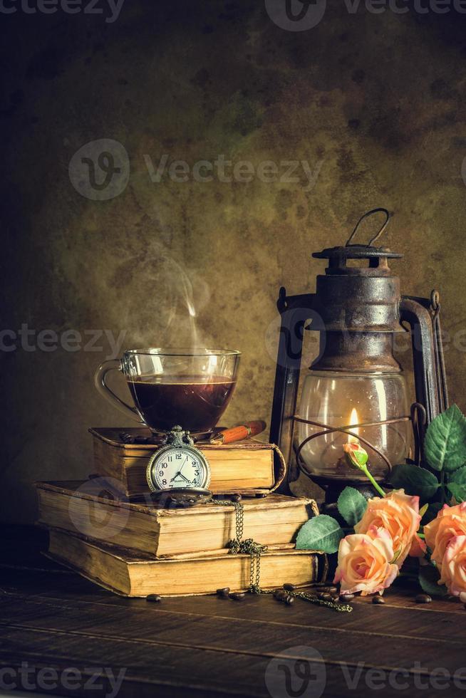 caffè in tazza di vetro su vecchi libri e orologio vintage con lampada a cherosene lanterna a olio che brucia con luce soffusa bagliore su pavimento in legno invecchiato. foto