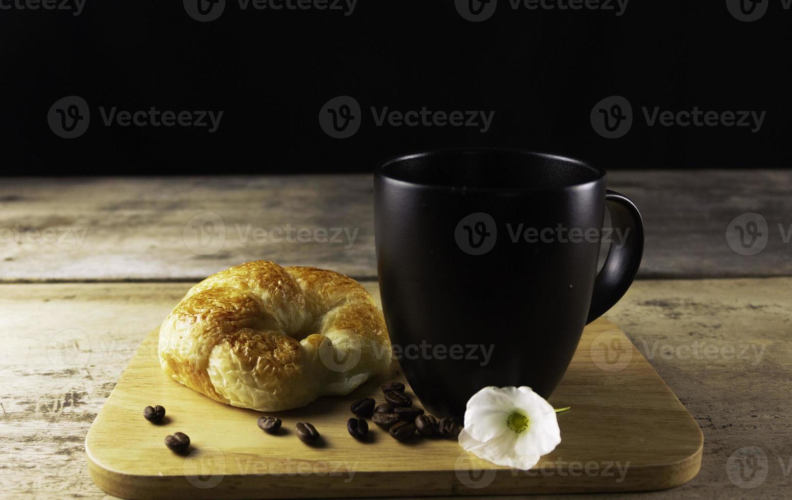 tazza di caffè con croissant, mucchio di chicchi di caffè tostati su un vecchio tavolo in legno rustico sfondo nero. girato in studio. foto