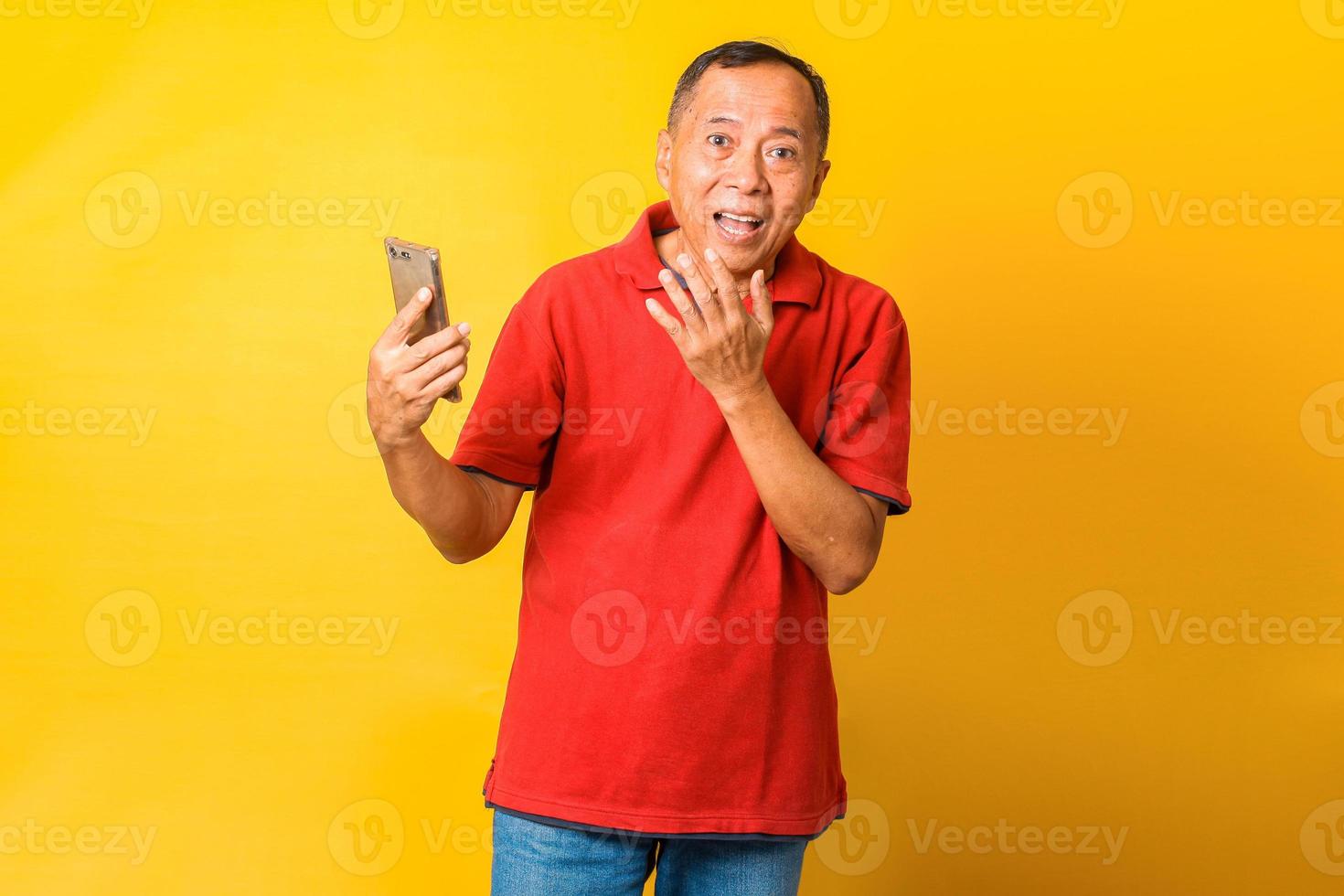 foto del nonno asiatico tenere telefono e bocca aperta sorpreso indossare maglietta rossa isolato colore giallo sfondo.