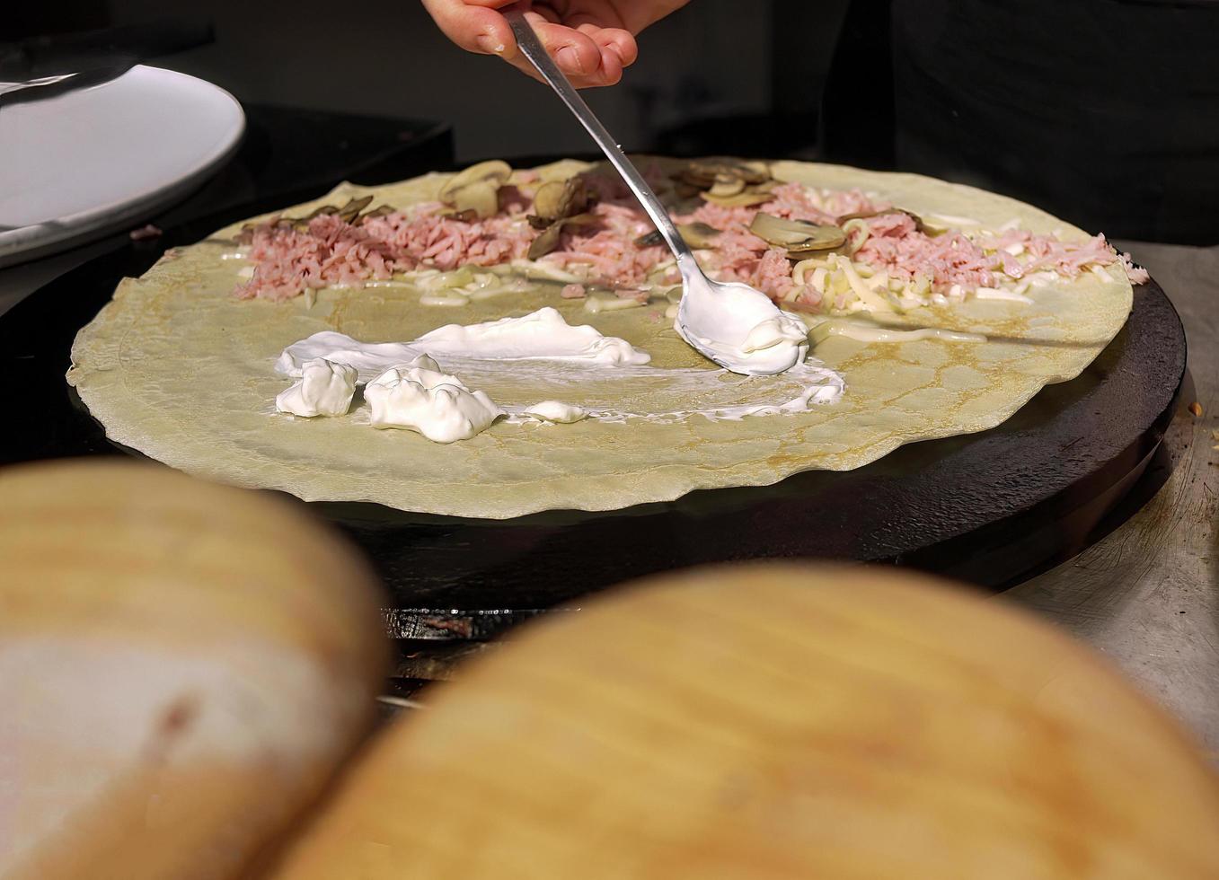 primo piano una mano che tiene un cucchiaio spalmando la crema sul prosciutto e sulla crepe ai funghi foto