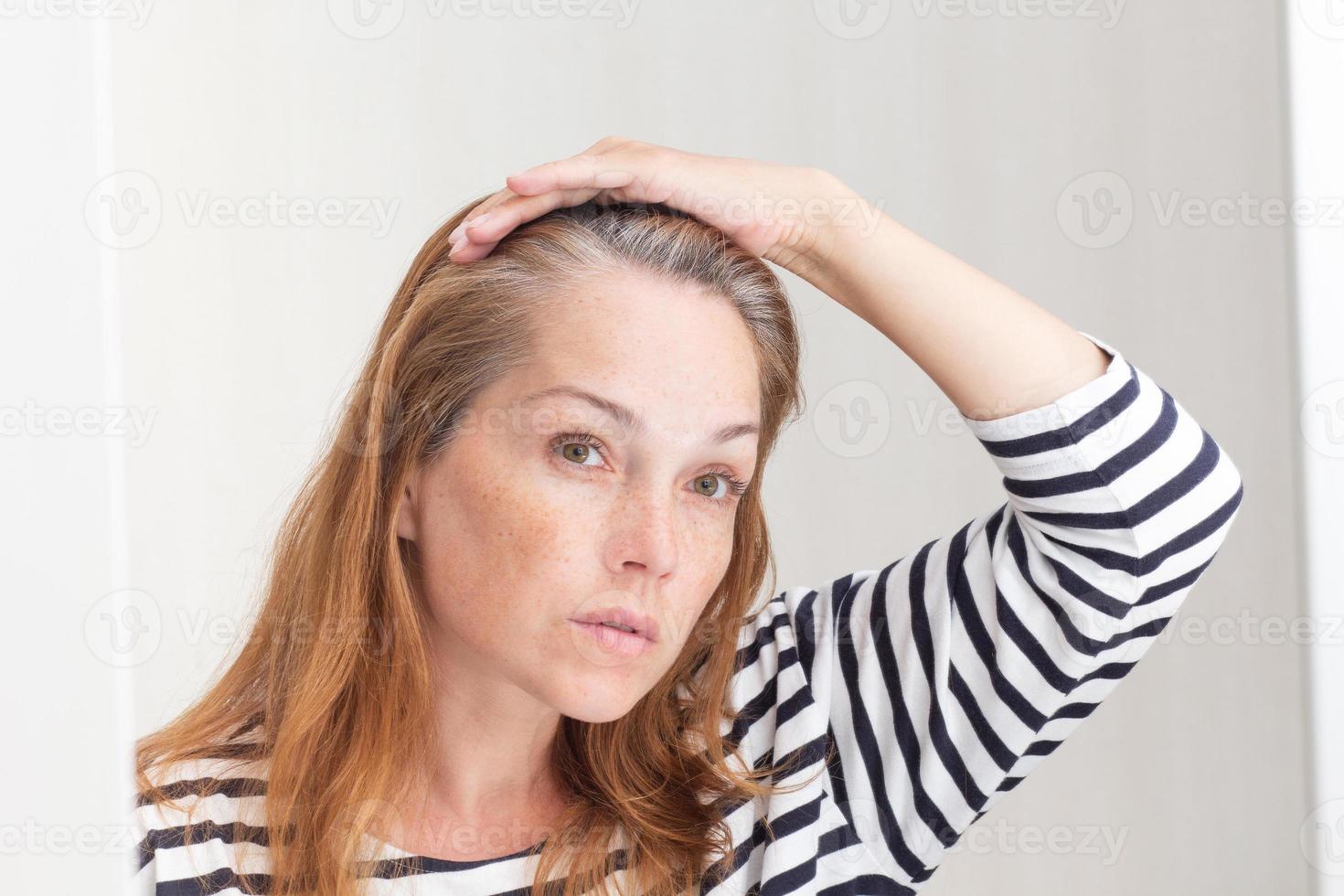 donna seria che guarda allo specchio i capelli grigi sulla radice di crescita foto