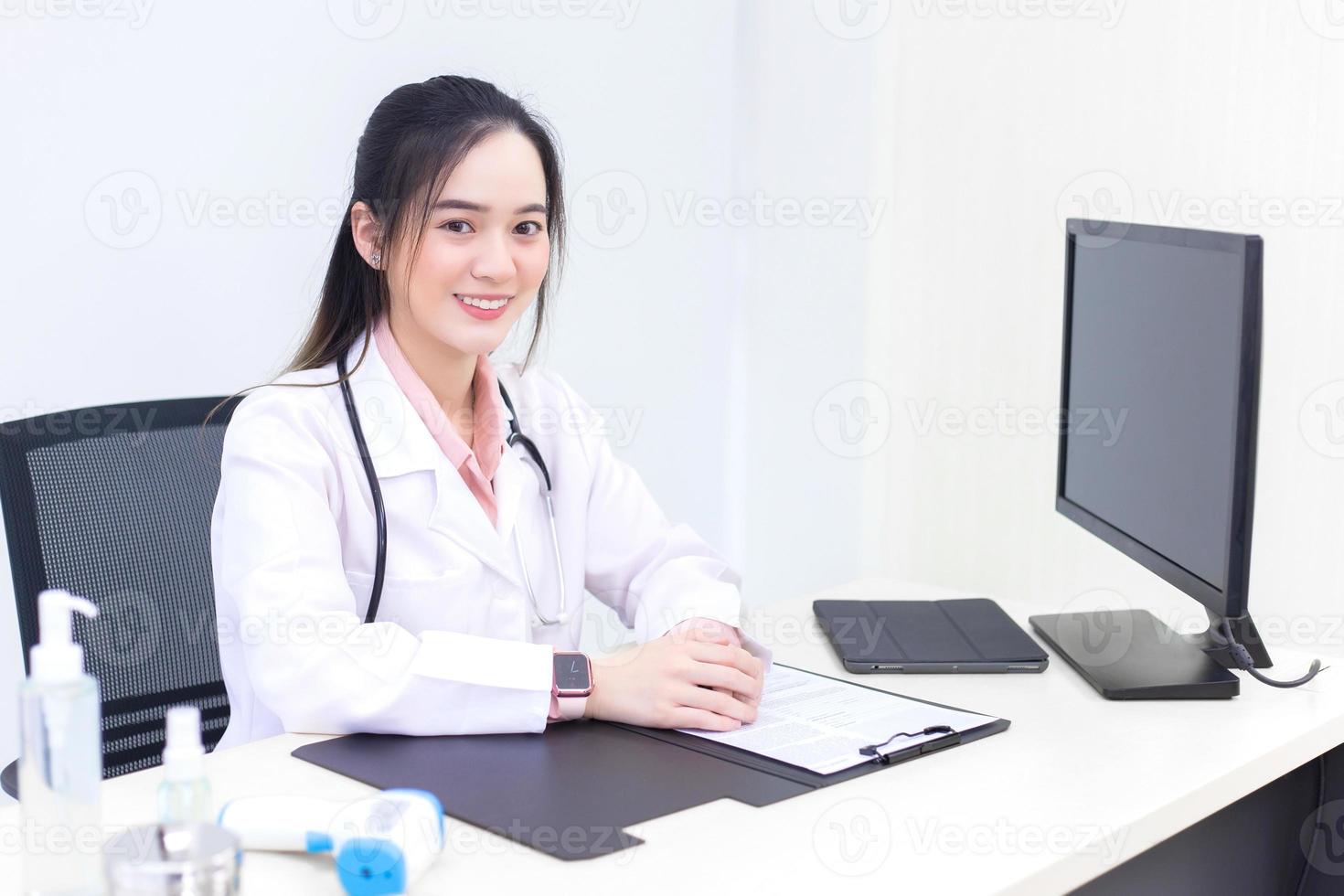 il medico asiatico della giovane donna in un camice bianco è seduto nella stanza dell'ufficio dell'ospedale foto