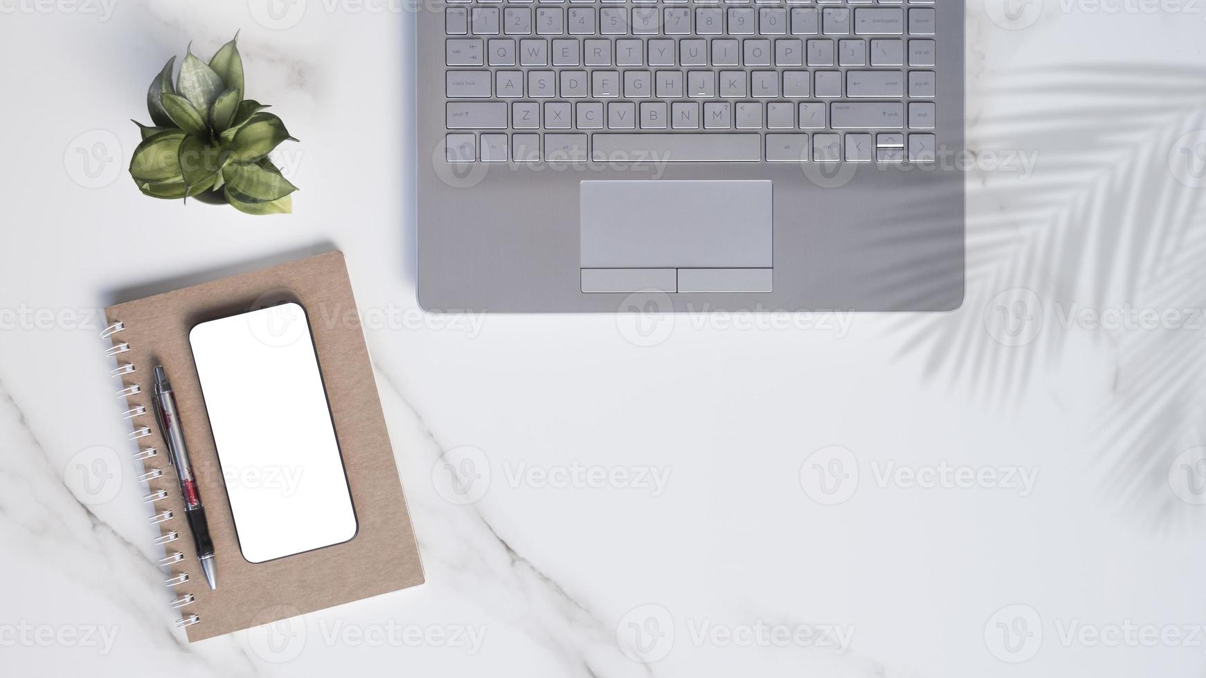 disposizione piatta del moderno tavolo da ufficio in marmo bianco con laptop, smartphone mock up, penna a sfera, taccuino a spirale con piccola pianta d'appartamento verde e ombra di foglie di palma sul tavolo foto