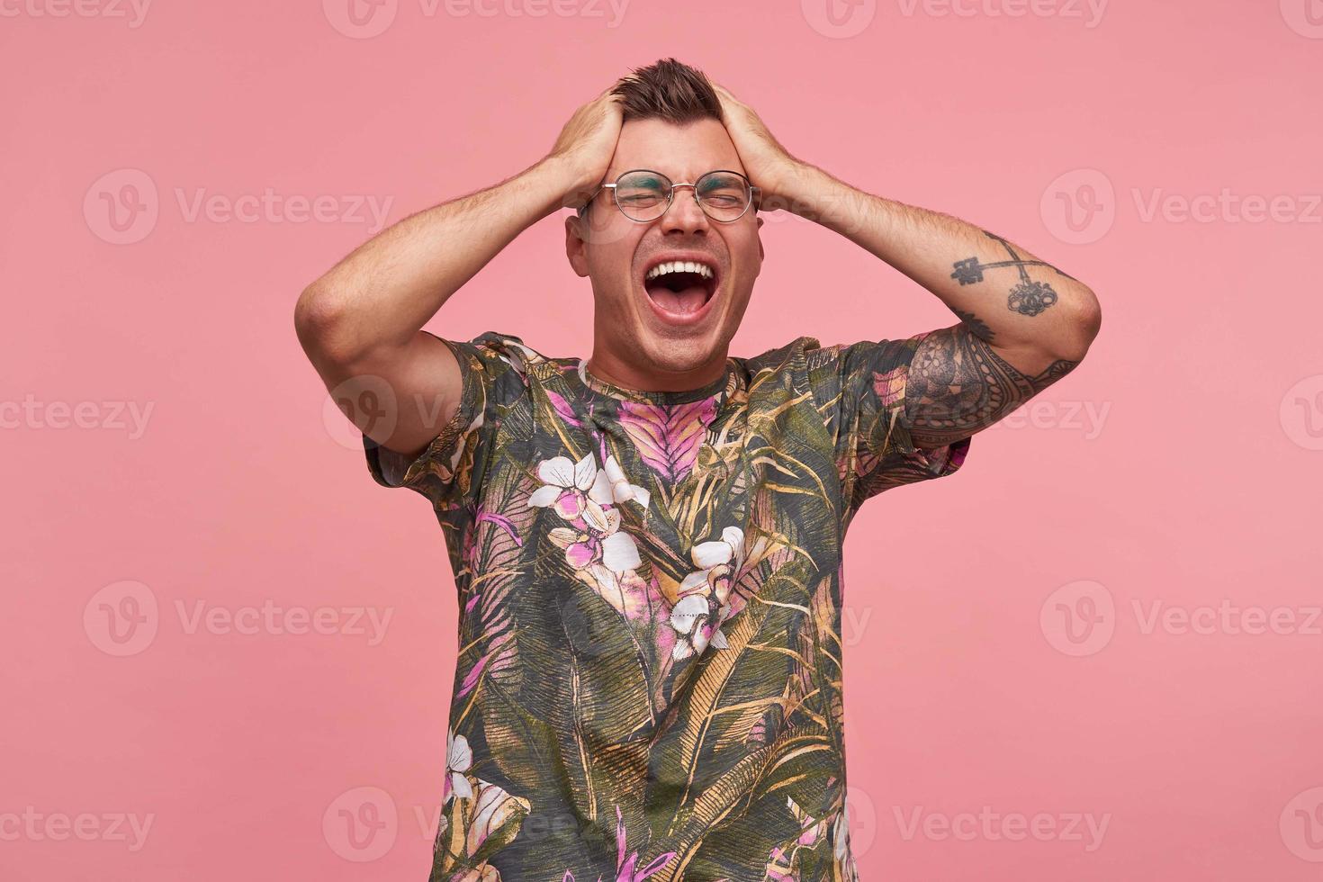 ragazzo attraente eccitato con tatuaggi in t-shirt casual in posa su sfondo rosa, in piedi con i palmi delle mani sulla testa, chiudendo gli occhi e gridando forte foto