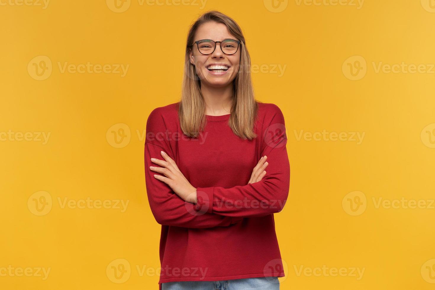 bella giovane ragazza bionda sorridente, che sembra felice alla telecamera, indossa un maglione rosso e occhiali, in piedi isolato su uno sfondo giallo foto