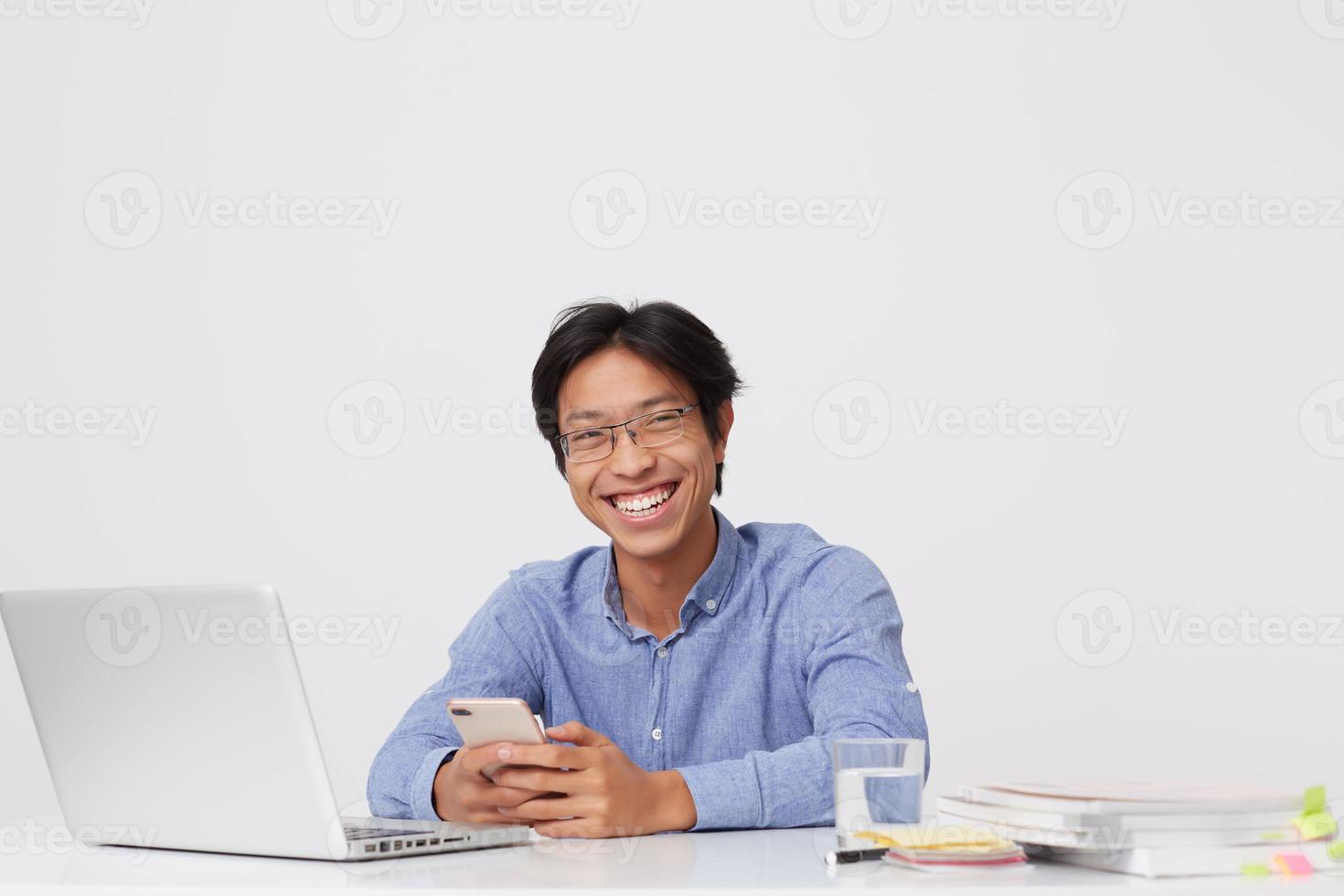 felice sorridente asiatico giovane uomo d'affari in bicchieri utilizzando il telefono cellulare ridendo e lavorando al tavolo con il computer portatile su sfondo bianco foto