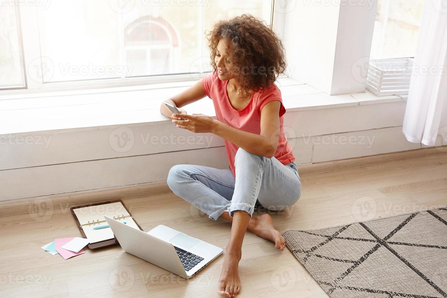 giovane e graziosa freelance che lavora a distanza da casa utilizzando un laptop moderno, cercando il numero di telefono nel notebook e andando a fare una chiamata con il suo smartphone foto