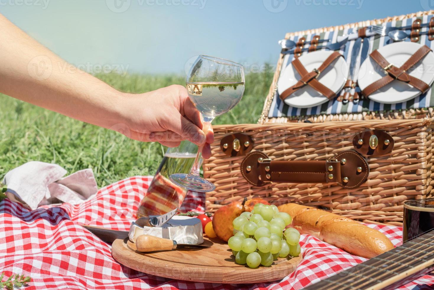 primo piano del cestino da picnic con bevande e cibo sull'erba. bicchiere di vino della holding della mano maschio foto