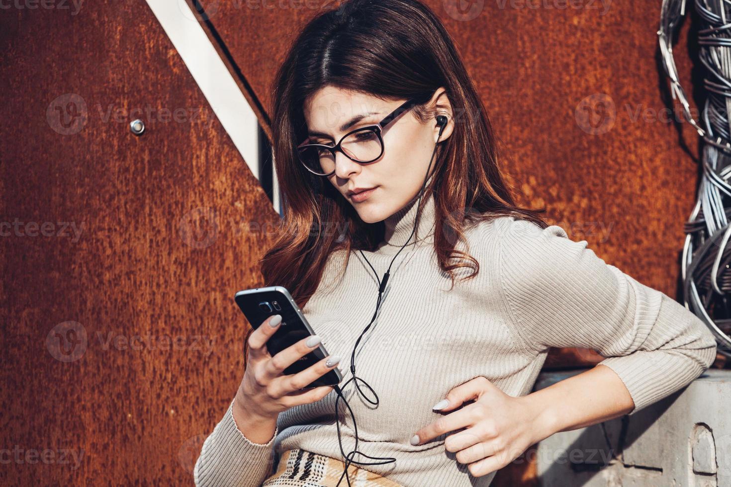 immagine di bella donna alla moda che tiene il telefono cellulare. giovane ragazza europea in piedi per strada e utilizzando il cellulare. donna che ascolta la musica con il telefono e si diverte. concetto di Internet mobile. foto