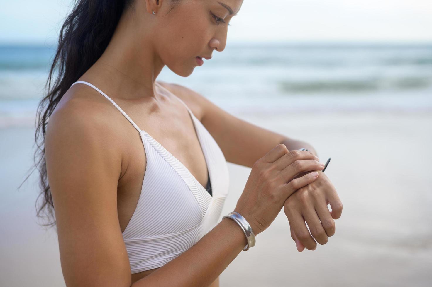 giovane bella donna in bikini rilassante e utilizzando smartwatch sulla spiaggia, estate, vacanze, vacanze e concetto di salute. foto