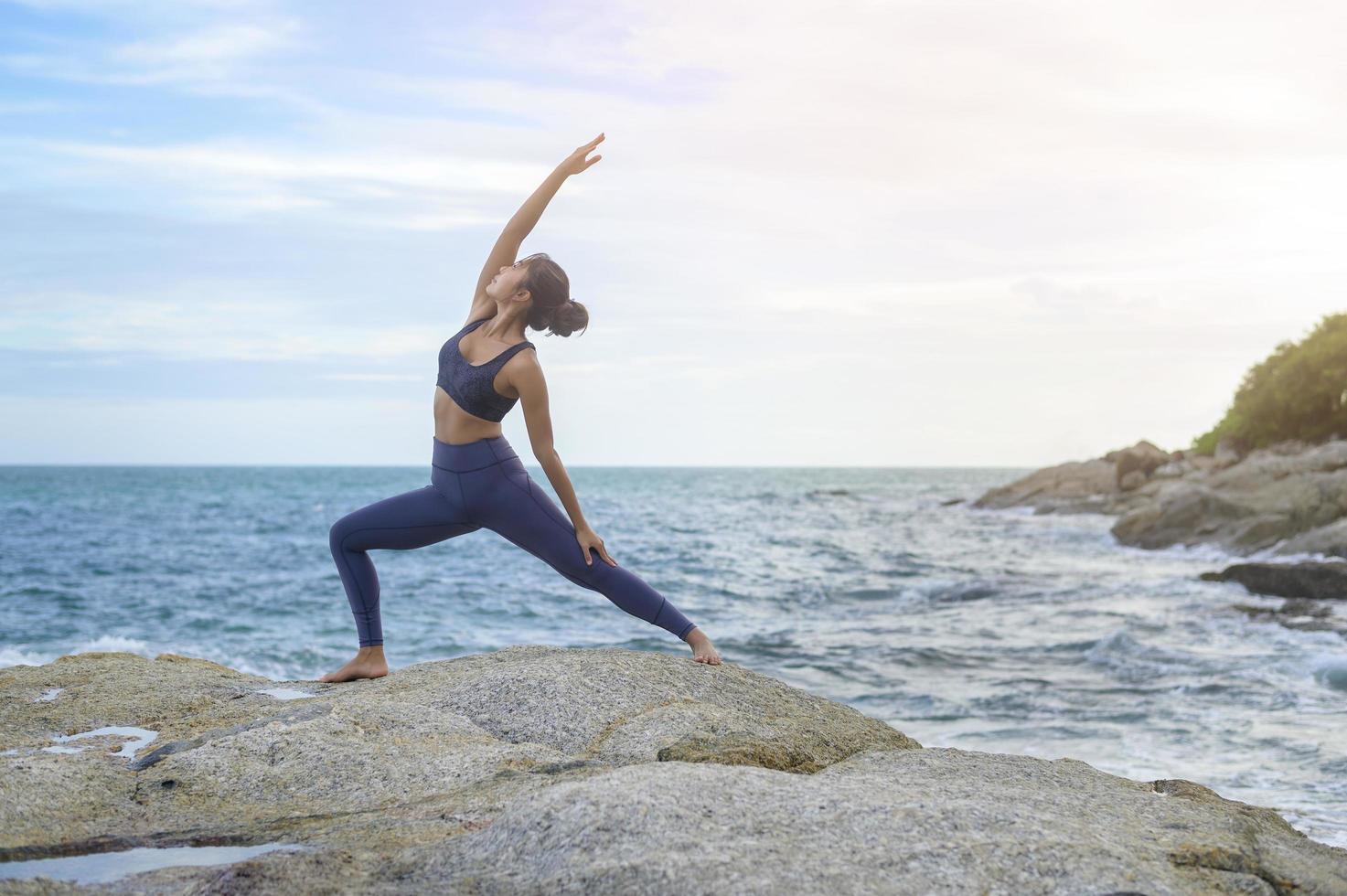 giovane donna asiatica in abbigliamento sportivo che fa yoga sulla roccia al mare, concetto di salute e meditazione foto