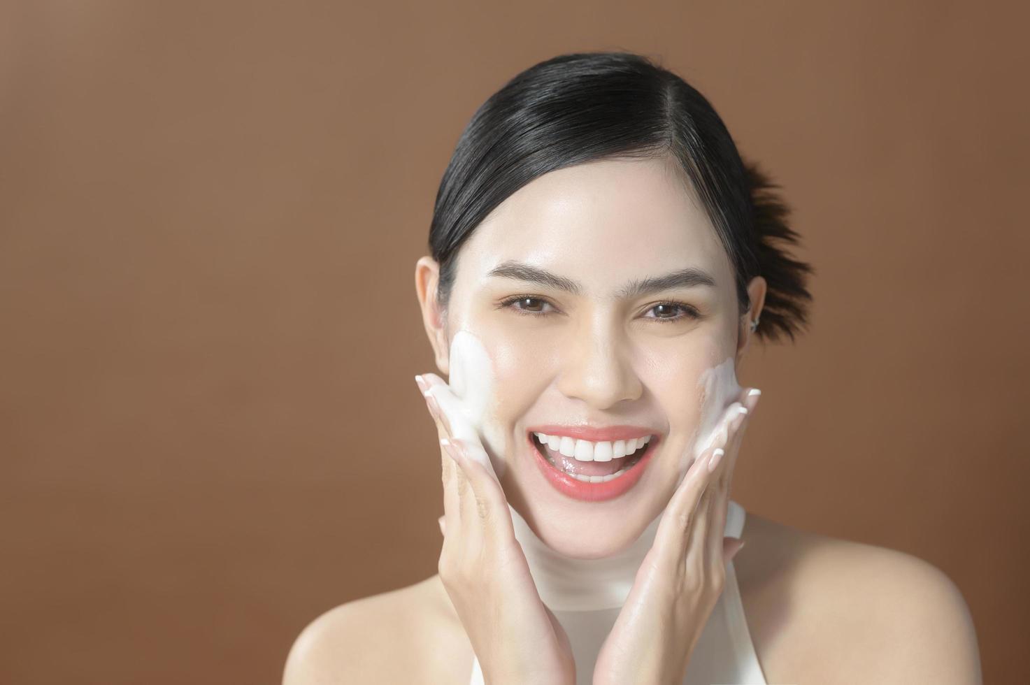 una giovane donna con un bel viso sorridente, lavandosi il viso su sfondo marrone, concetto di bellezza per la cura della pelle foto