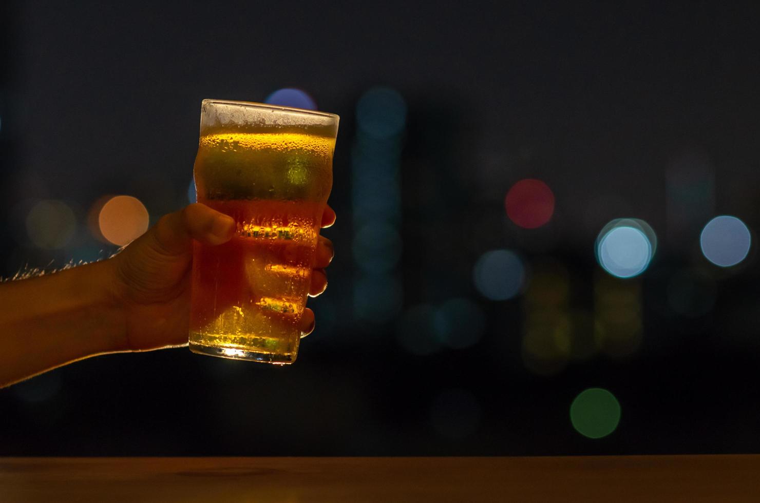 mano con un bicchiere di birra che brinda per la celebrazione e il concetto di festa isolato su sfondo notturno scuro con luci colorate del bokeh della città sul bar sul tetto. foto