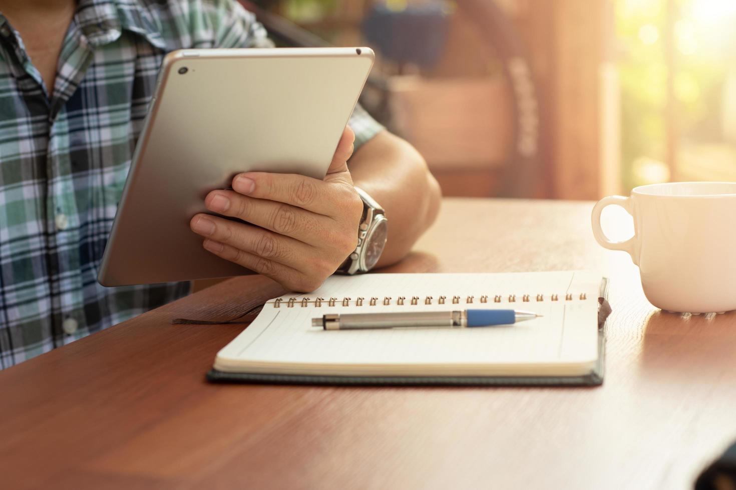 il giovane si siede a casa a tavola, usa la tavoletta digitale su scrivania e taccuino, tazza di caffè. uomo che lavora, studia. foto