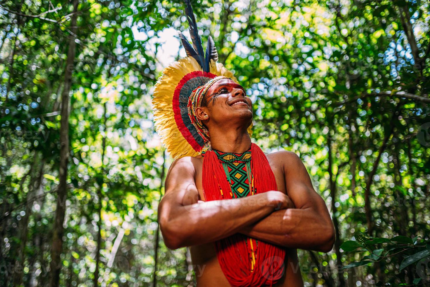 indiano della tribù pataxo, con copricapo di piume. giovane indiano brasiliano che guarda a destra, sorridente e le braccia incrociate foto