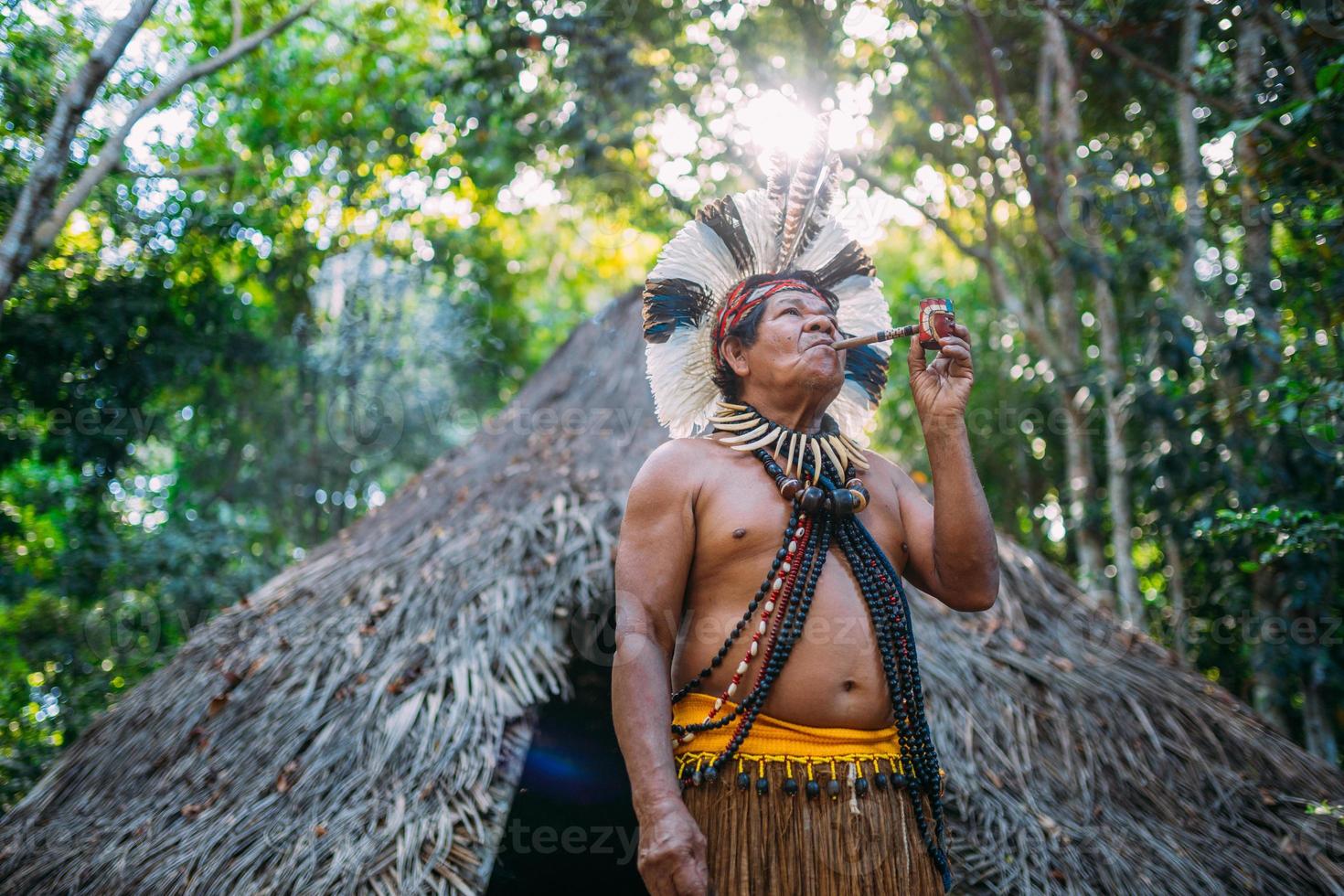 sciamano della tribù pataxo, che indossa un copricapo di piume e fuma la pipa. indiano brasiliano guardando a destra foto