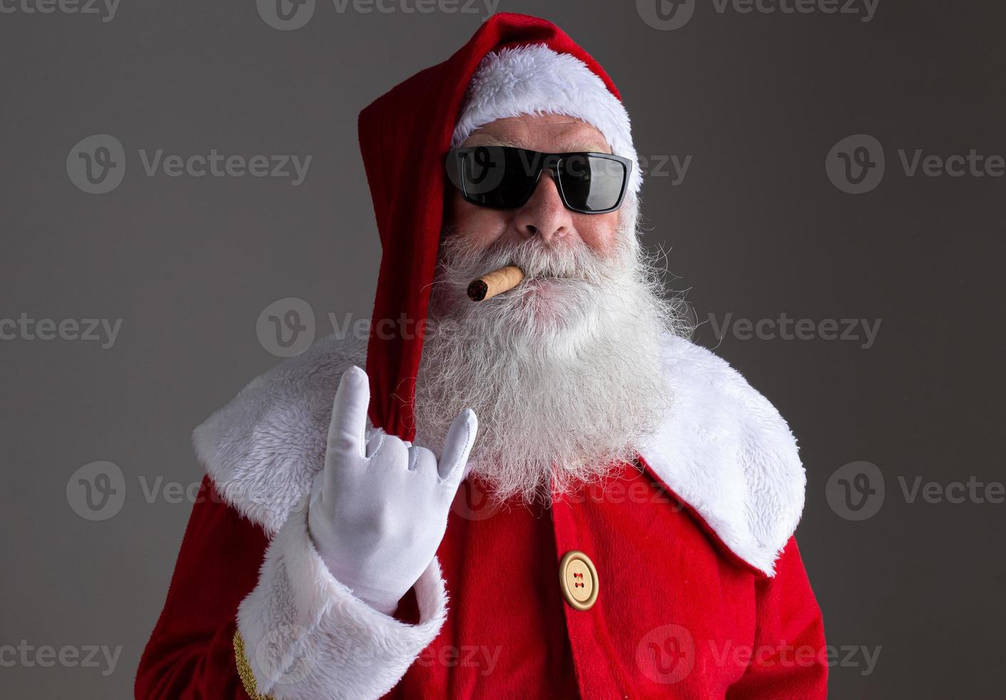 Babbo Natale che indossa occhiali da sole che mostra il segno della mano del bilanciere e fuma un sigaro su sfondo scuro foto
