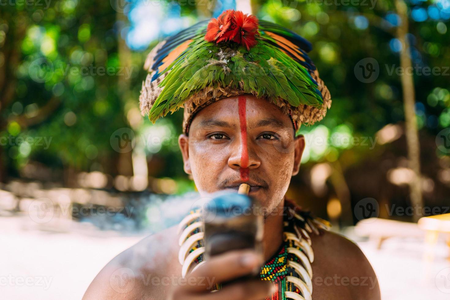 capo indiano della tribù pataxo che fuma la pipa . indiano brasiliano con copricapo di piume e collana che guarda l'obbiettivo foto