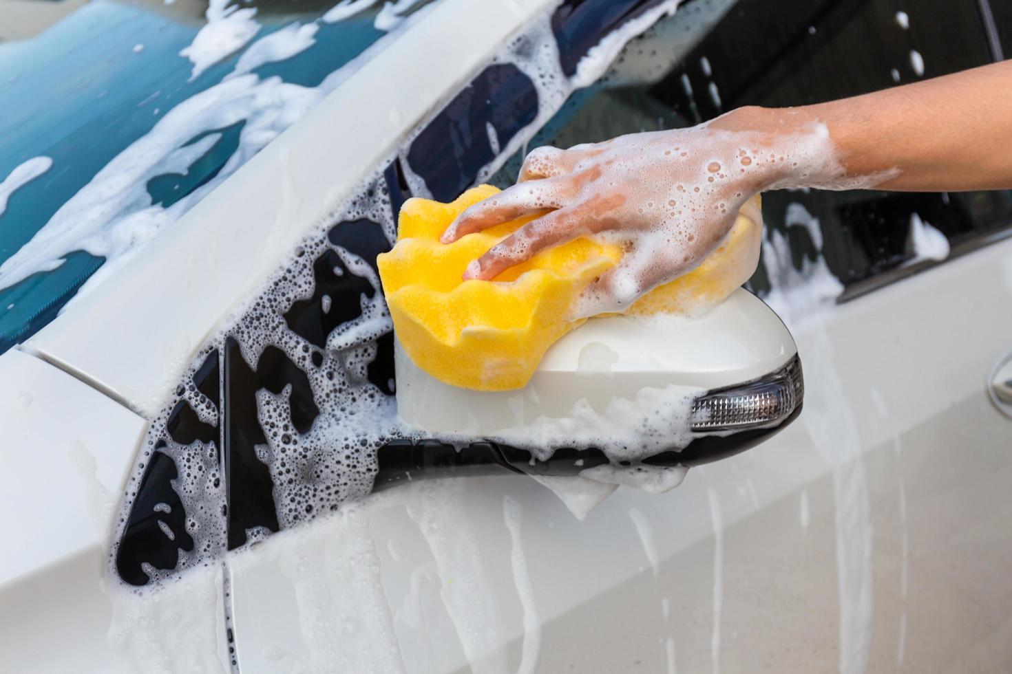 mano della donna con la spugna gialla che lava lo specchio laterale dell'automobile moderna o dell'automobile di pulizia. concetto di autolavaggio foto