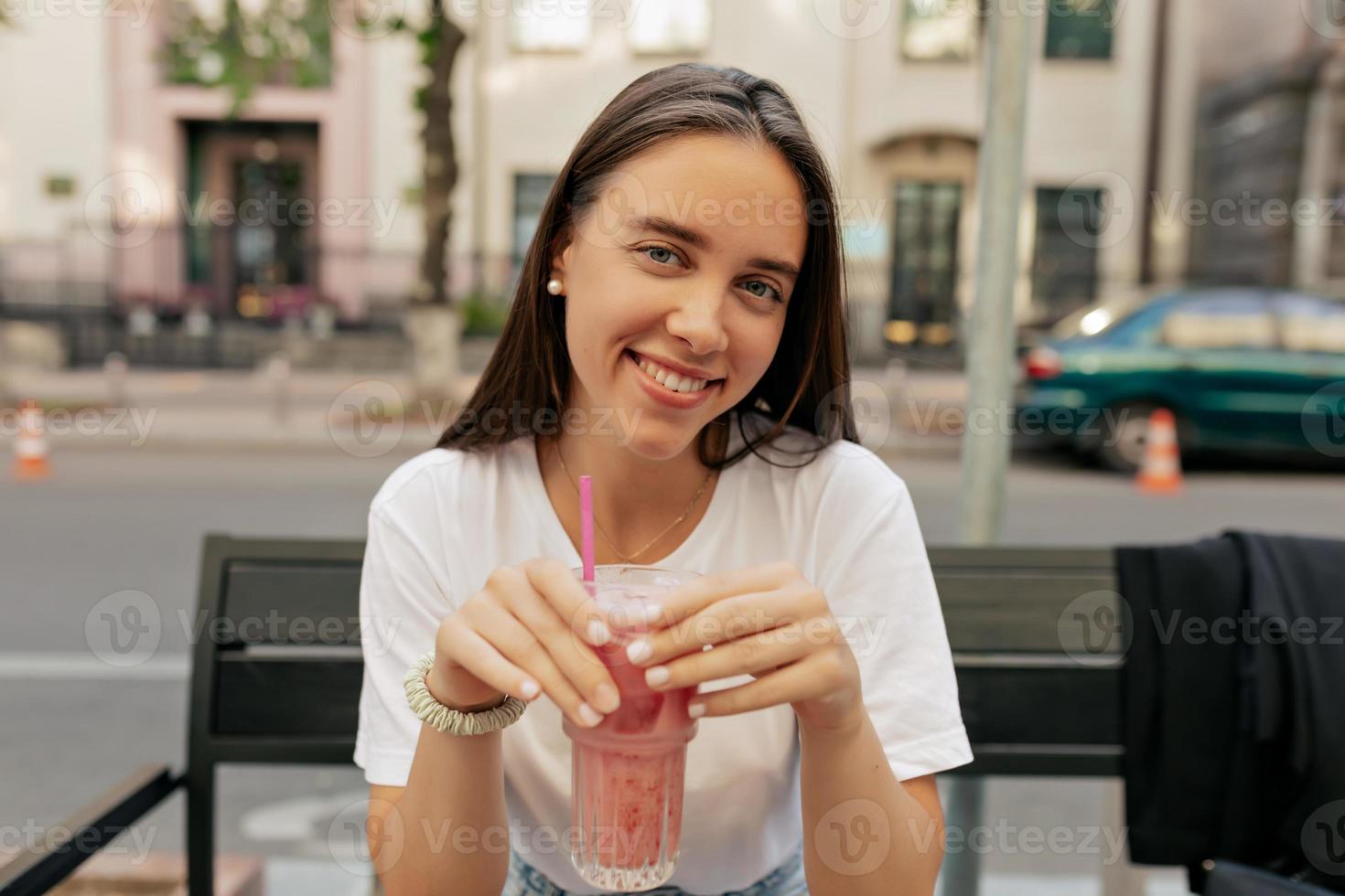 Ritratto ravvicinato di adorabile donna sorridente con un bel sorriso e capelli castani che beve frullato e posa davanti alla telecamera nella caffetteria all'aperto foto