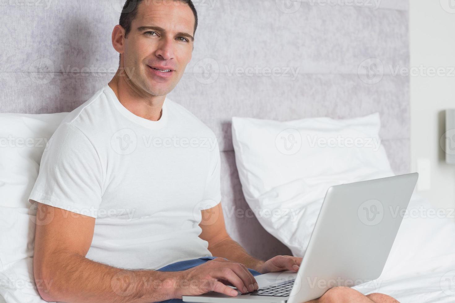 uomo allegro che per mezzo del computer portatile sul letto foto