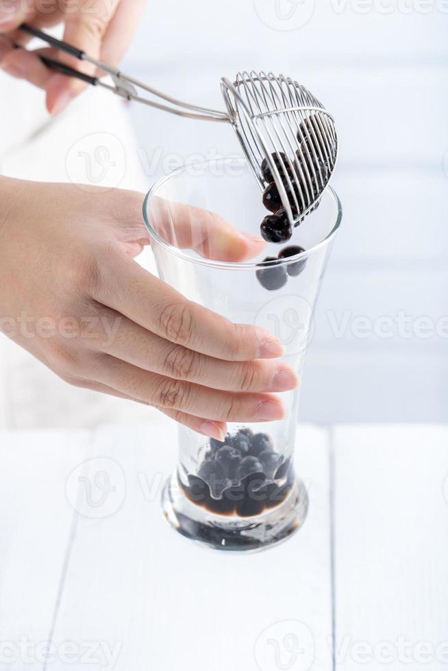 preparare il tè a bolle, scoop e versare le palline di bolla di perle di tapioca al gusto di zucchero di canna cotto nella tazza su sfondo bianco del tavolo in legno, primo piano, spazio per la copia. foto