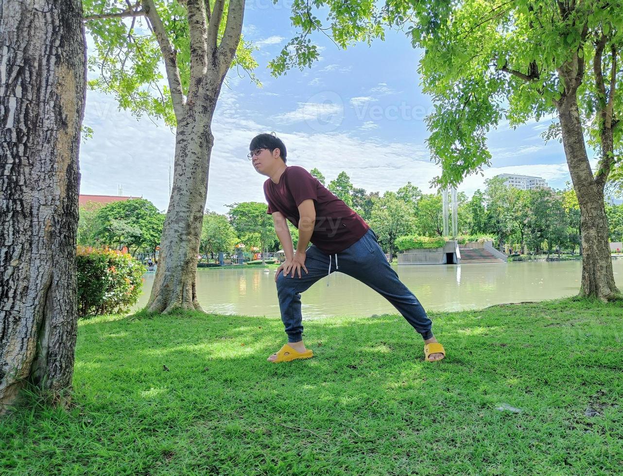 giovane uomo asiatico che indossa occhiali con capelli scuri in piedi yoga o riscaldamento sotto un albero. nella città del parco estivo foto