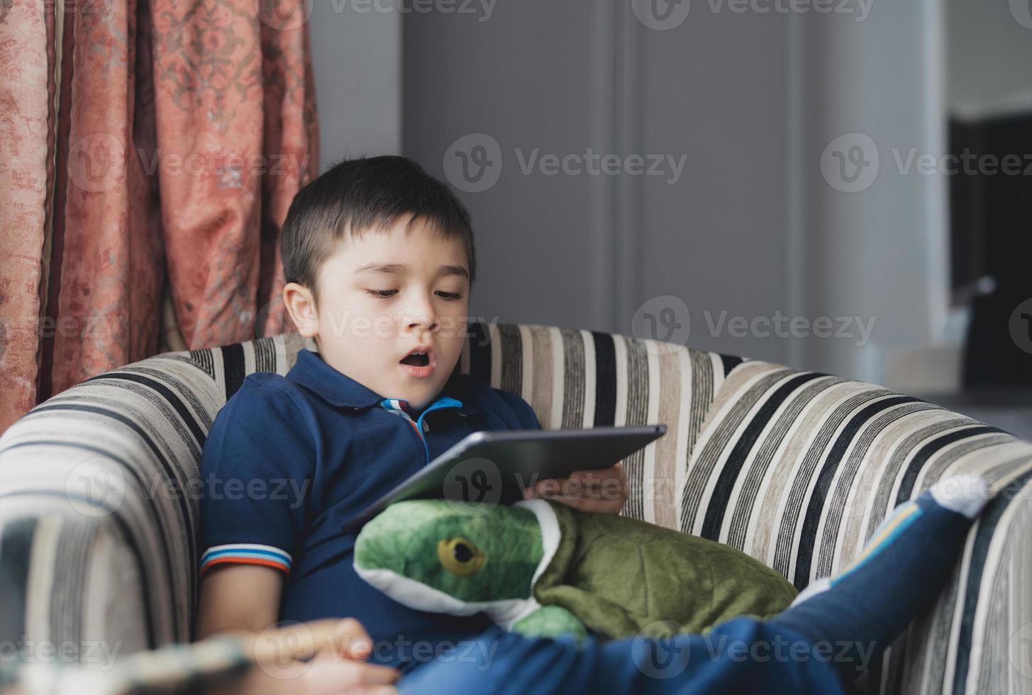 ritratto cinematografico ragazzo che gioca sul tablet seduto sul divano con la luce che splende dalla finestra, bambino che gioca online su internet a casa, bambino che parla videochiamata con gli amici a casa foto