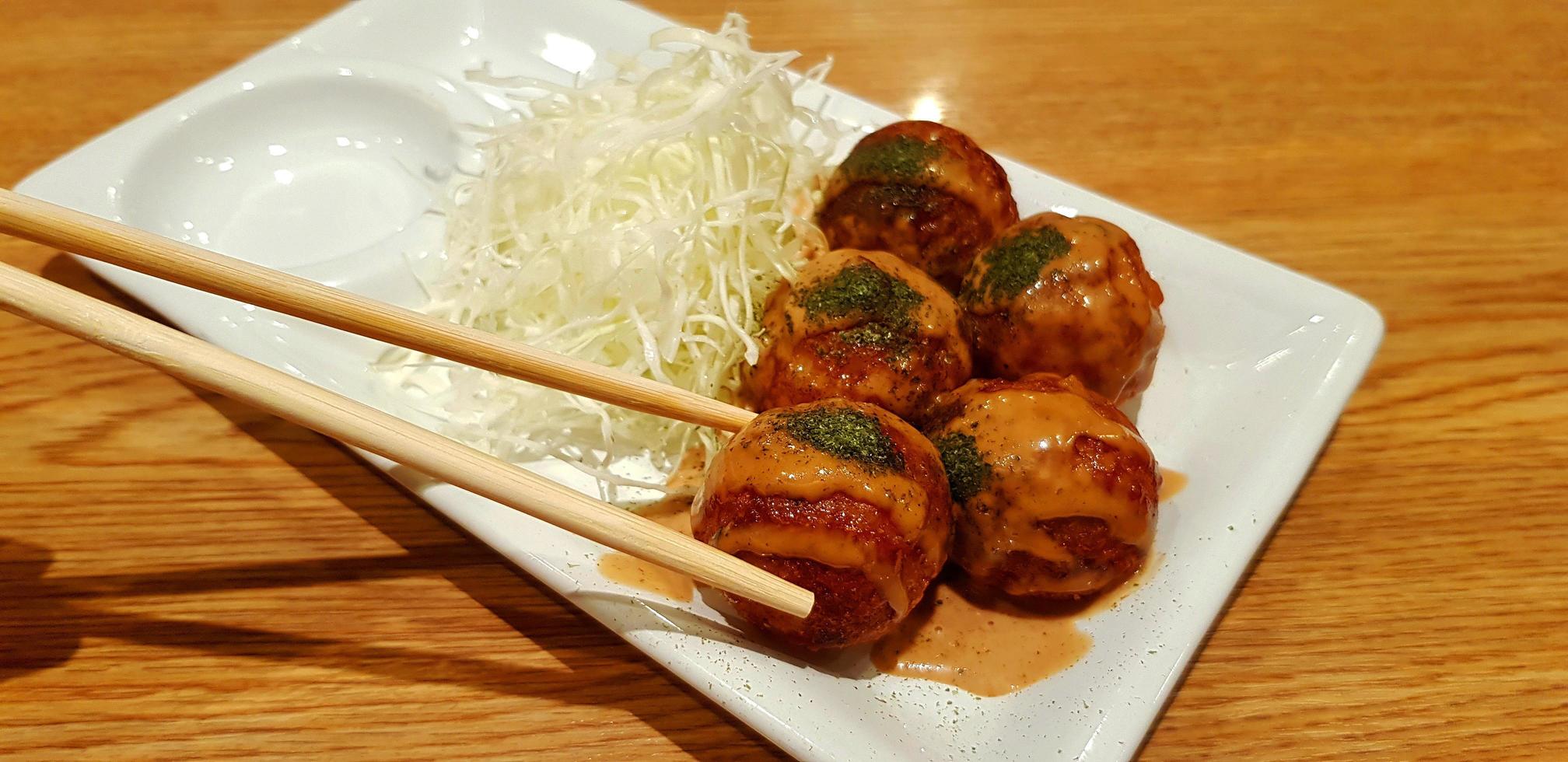 takoyaki condito con salsa dolce e cavolo affettato in un piatto bianco su fondo di legno marrone. mangiare cibo giapponese utilizzando le bacchette nel piatto su un tavolo di legno con spazio per la copia. foto