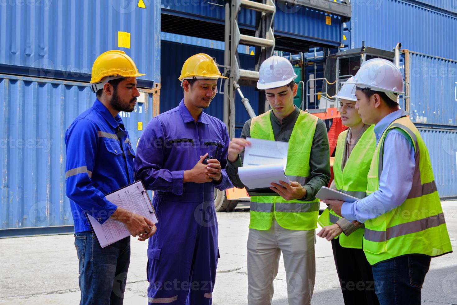 un gruppo di lavoratori multirazziali in divisa di sicurezza ed elmetti protettivi lavorano al terminal logistico con molte pile di container, caricando merci di spedizione controllate per l'industria del trasporto merci. foto