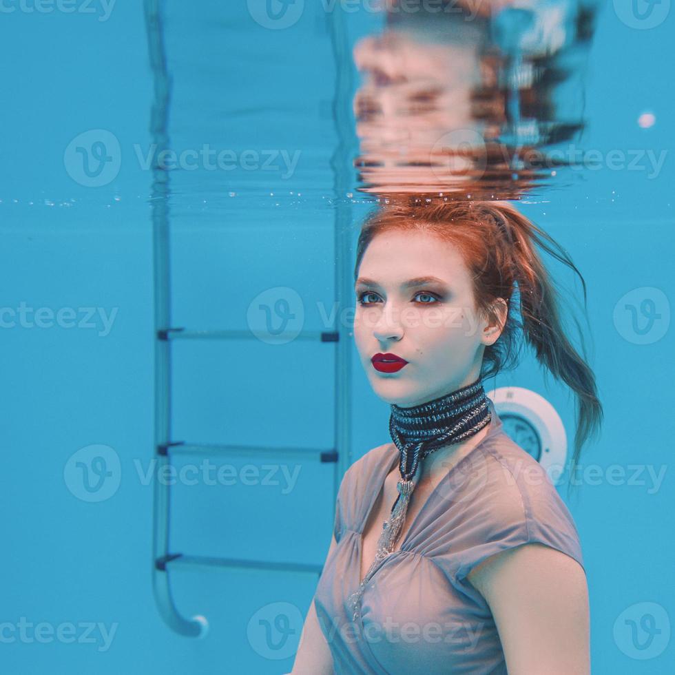 ritratto di arte surreale di giovane donna in abito grigio e sciarpa di perline sott'acqua in piscina foto