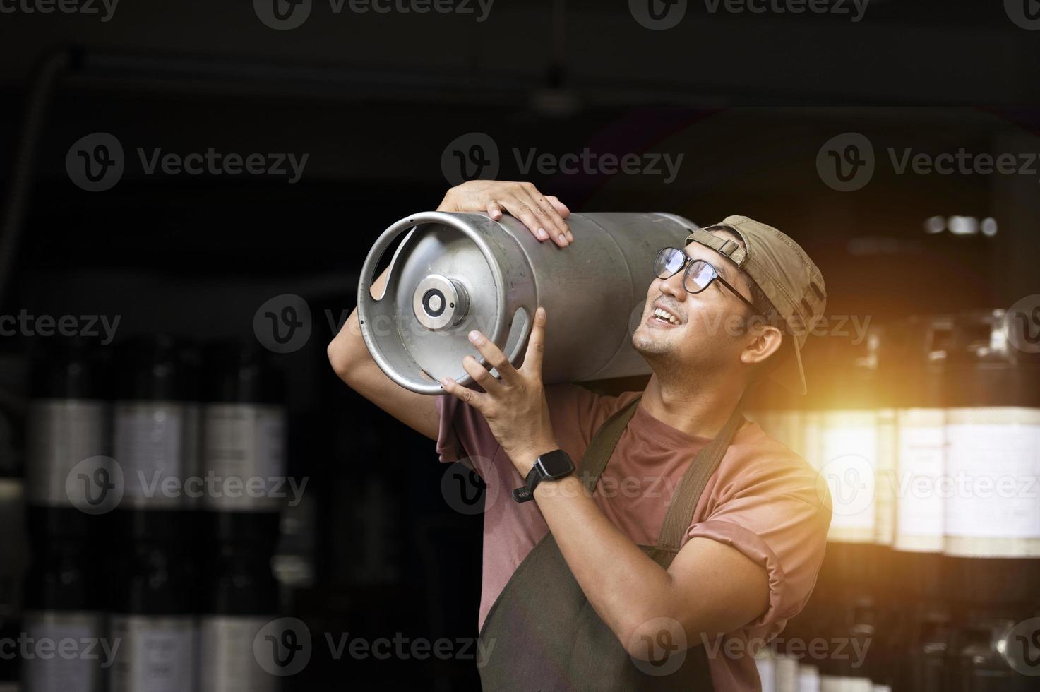 giovane uomo in grembiule di pelle che tiene un fusto di birra al moderno birrificio, operaio del birrificio artigianale foto