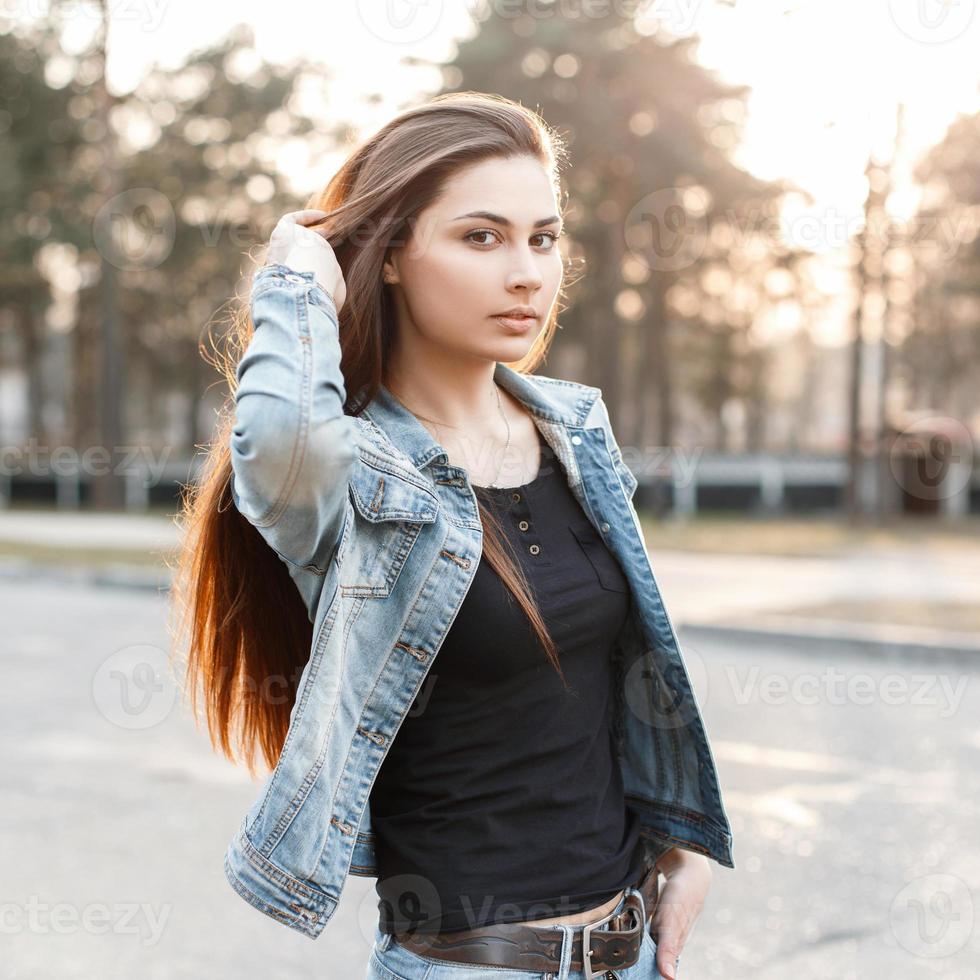 la ragazza alla moda in vestiti alla moda dei jeans corregge i suoi capelli lunghi al tramonto foto