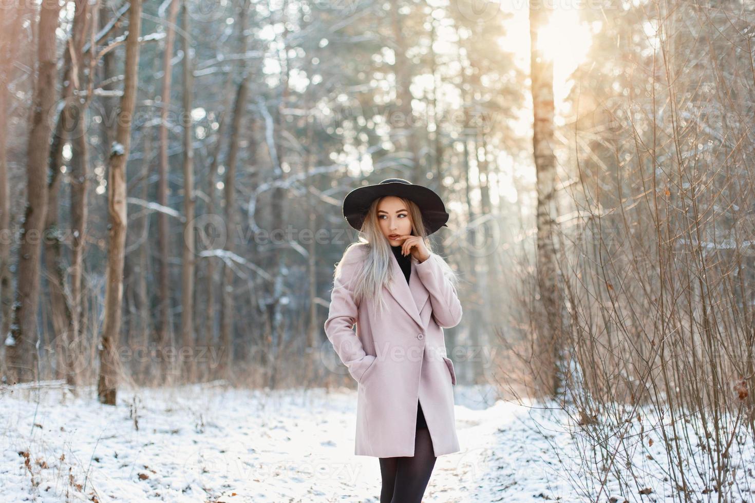 giovane donna alla moda con un cappello e un cappotto in piedi in una foresta invernale al tramonto foto