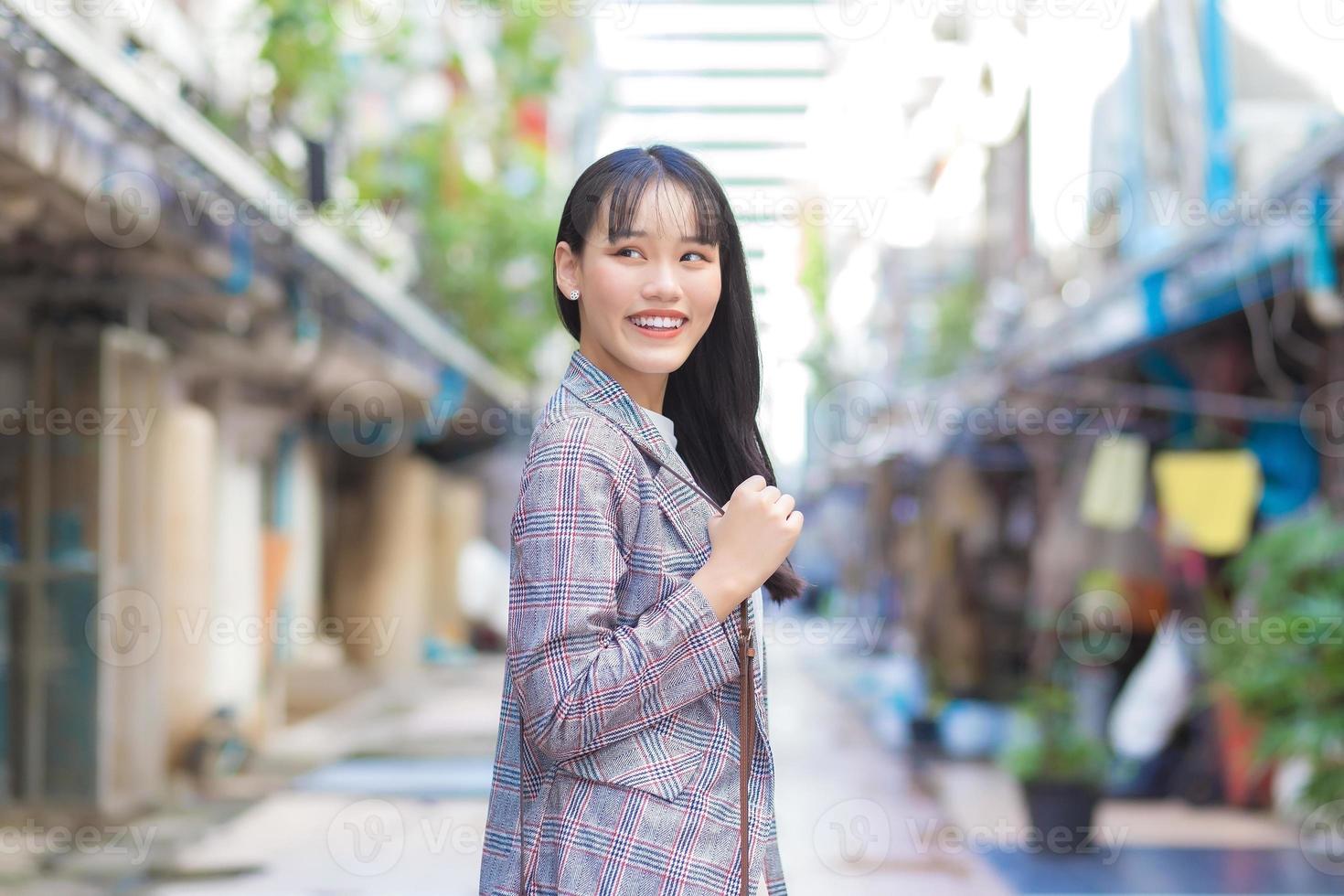 una giovane donna asiatica sicura di sé che indossa un blazer a righe marroni e una borsa a tracolla sorride felice mentre si reca al lavoro attraverso la città vecchia. foto