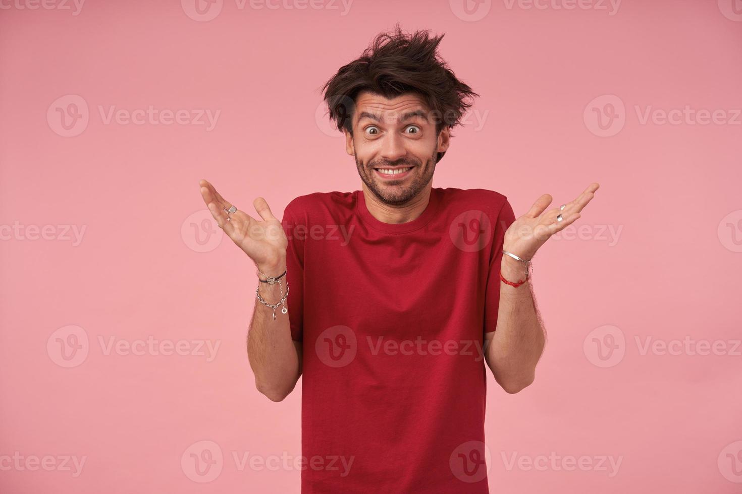 ritratto di giovane uomo con gli occhi aperti con i capelli selvaggi in piedi su sfondo rosa con i palmi sollevati, indossando abiti casual, guardando la fotocamera con un ampio sorriso e alzando le sopracciglia foto