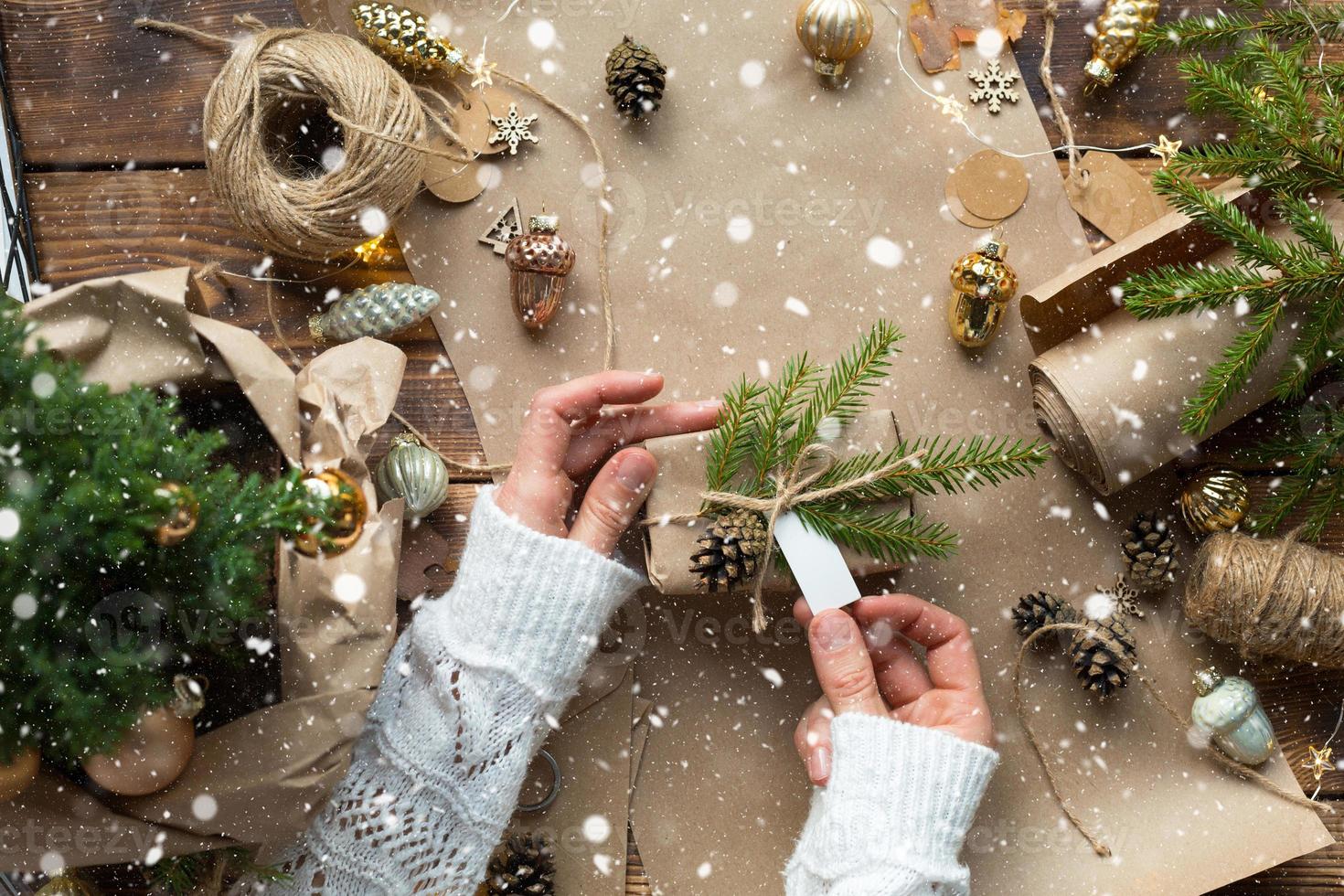 le mani confezionano un regalo per natale e capodanno in materiali ecologici carta kraft, rami di abete vivo, pigne, spago, legati con un fiocco. tag con mock up, arredamento naturale, fatto a mano, piatto. umore festivo foto