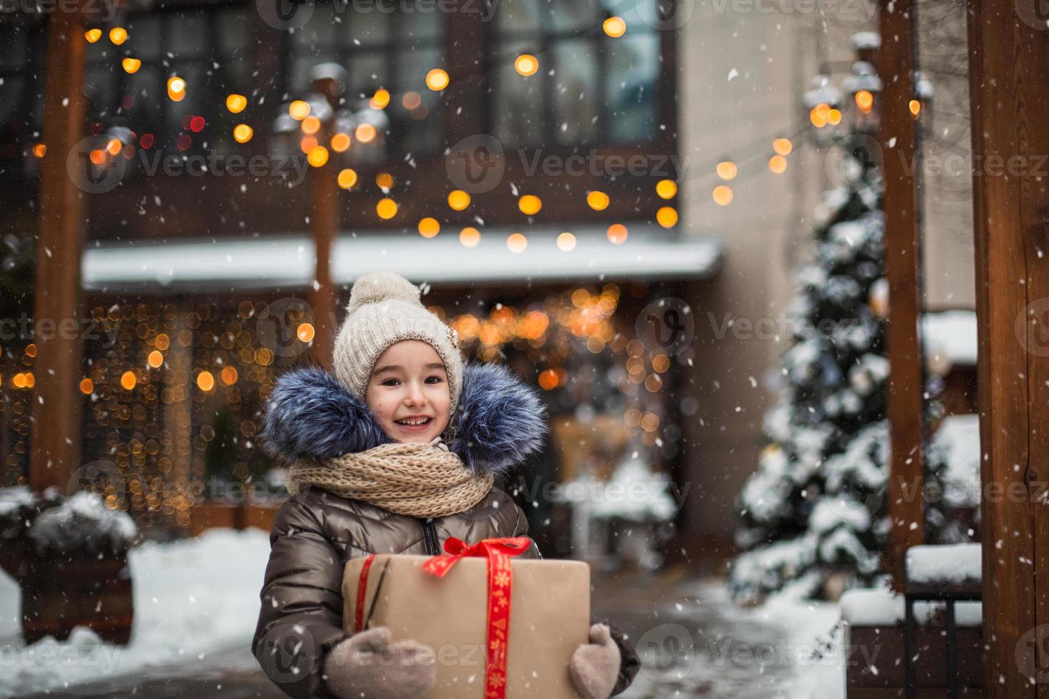 ritratto di ragazza gioiosa con una confezione regalo per natale in una strada cittadina in inverno con la neve in un mercato festivo con decorazioni e lucine. vestiti caldi, cappello lavorato a maglia, sciarpa e pelliccia. Capodanno foto