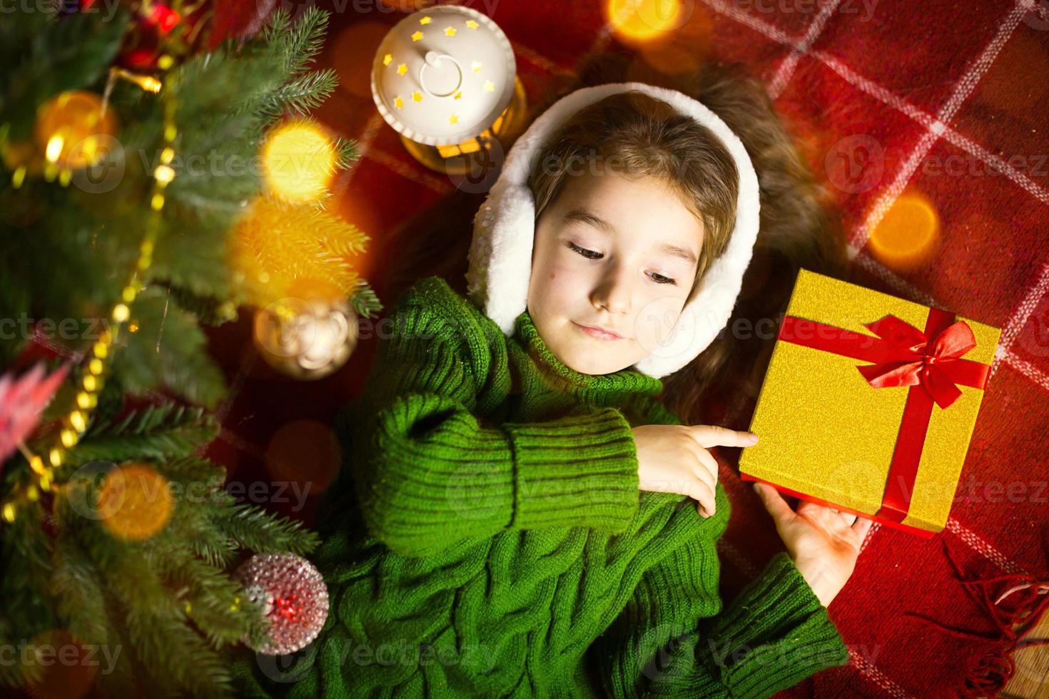 ragazza in un caldo maglione lavorato a maglia e cuffie di pelliccia è sdraiata su una coperta vicino all'albero di natale con una confezione regalo. vigilia di natale, un sogno e un desiderio. capodanno, atmosfera festiva, ghirlande e bokeh foto