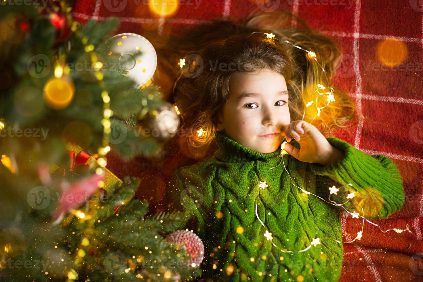 una ragazza con i capelli lunghi e le ghirlande giace su un plaid rosso sotto un albero di natale con i giocattoli in un caldo maglione lavorato a maglia. natale, capodanno, emozioni dei bambini, gioia, attesa di un miracolo e doni foto