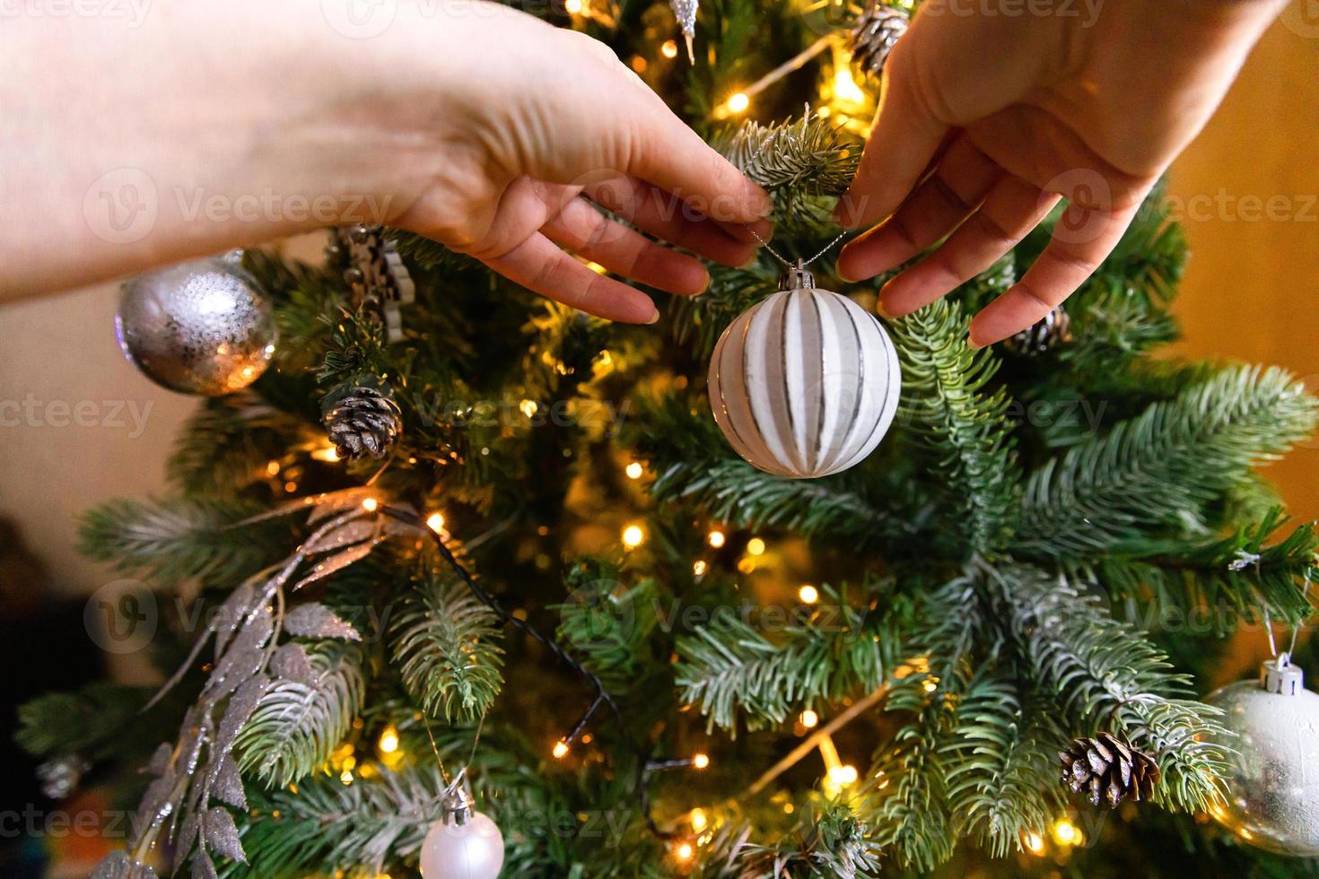 mani della donna che decorano l'albero di natale. albero di natale con decorazioni bianche e argento, ornamenti giocattolo e palla. design d'interni in stile classico moderno. la vigilia di natale a casa, tempo di festeggiamenti. foto