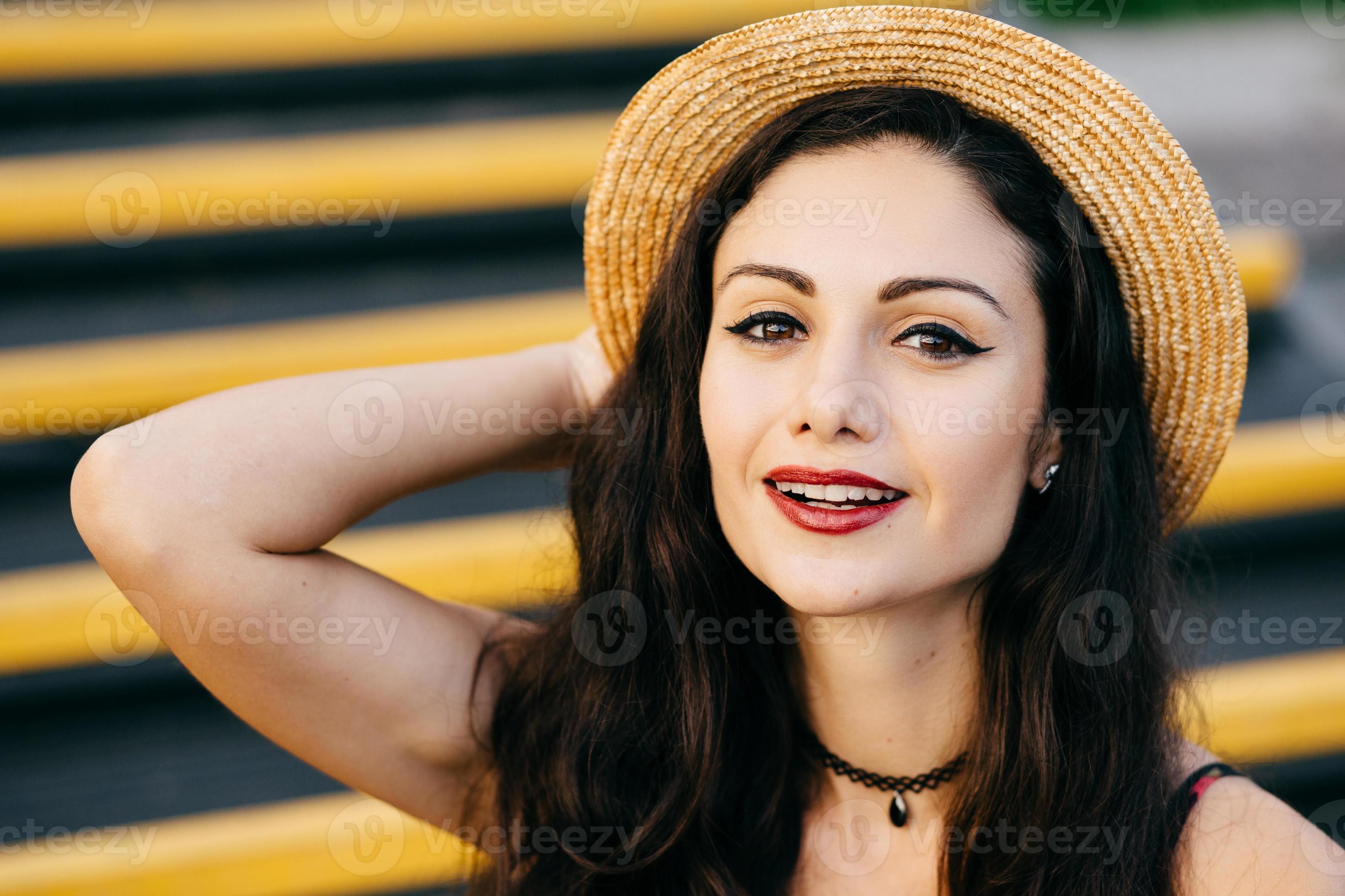 bella donna con i capelli scuri, occhi brillanti e labbra dipinte di rosso  che guarda con sicurezza e con un sorriso alla telecamera che indossa  cappello di paglia e collana sul collo.