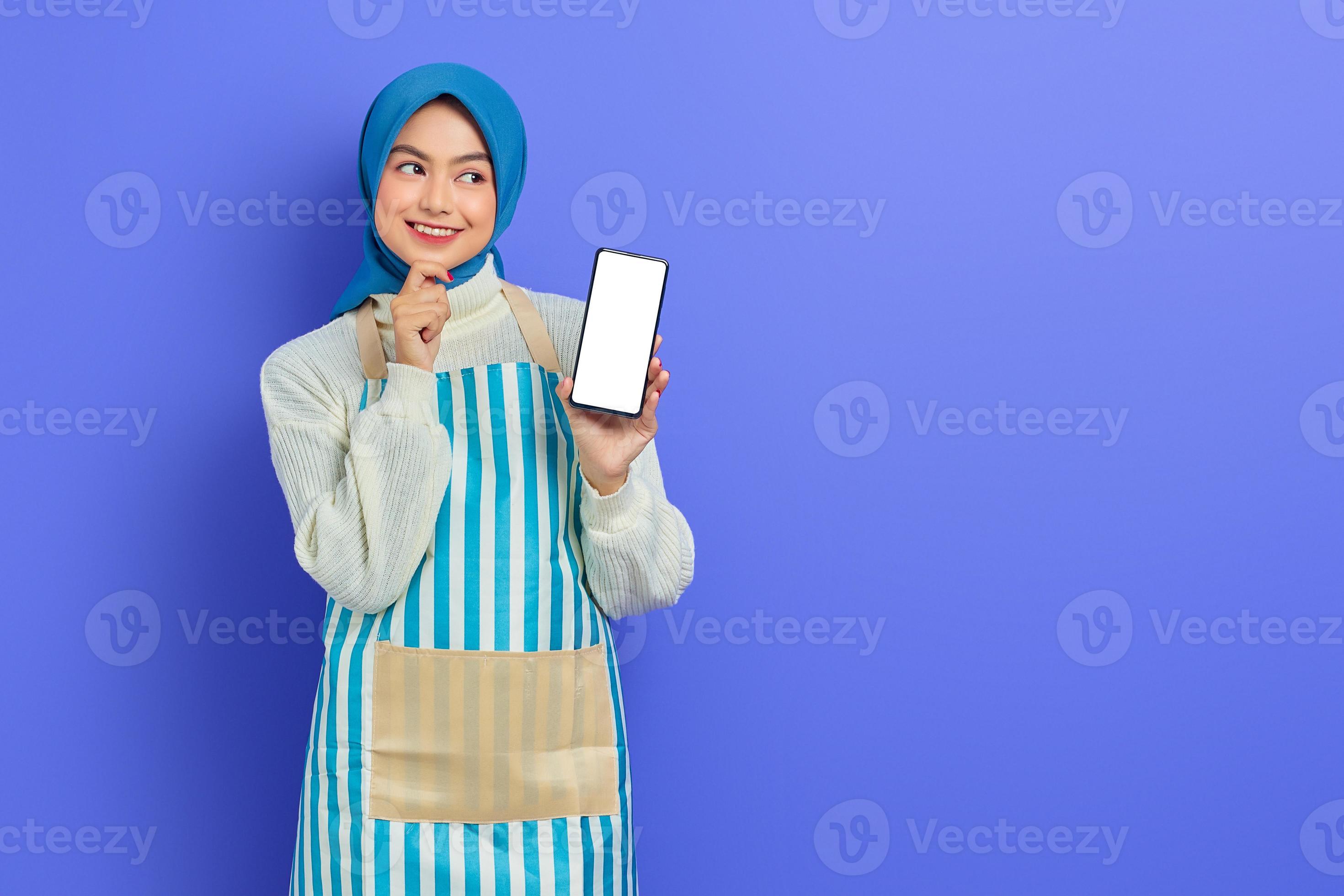 sorridente giovane donna musulmana asiatica che indossa hijab e grembiule che tiene il telefono cellulare con schermo vuoto mentre si tocca il mento, guardando da parte isolato su sfondo viola. concetto di stile di vita musulmano casalinga di persone foto