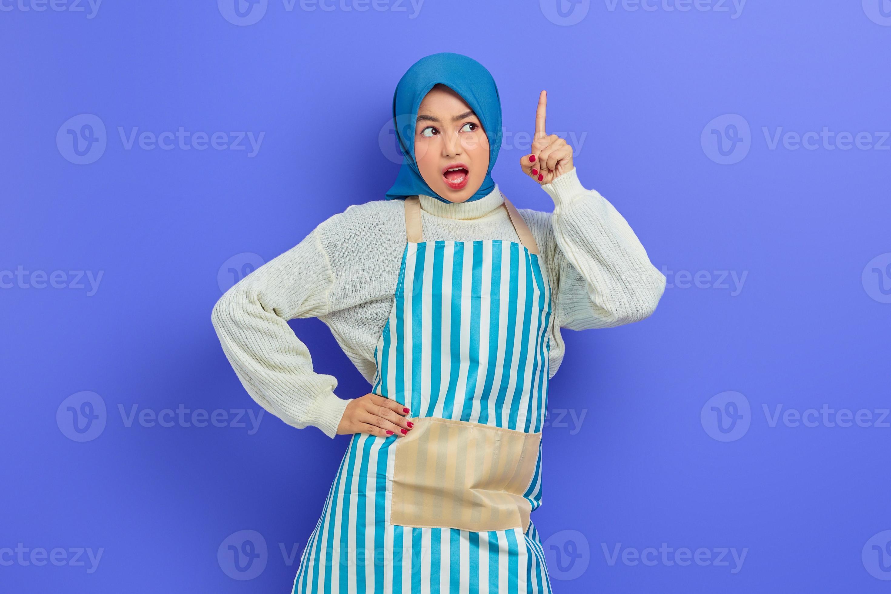 Ritratto di giovane donna musulmana asiatica scioccata in hijab e grembiule a righe, bocca aperta mentre indica lo spazio della copia con il dito isolato su sfondo viola. concetto di stile di vita musulmano casalinga di persone foto