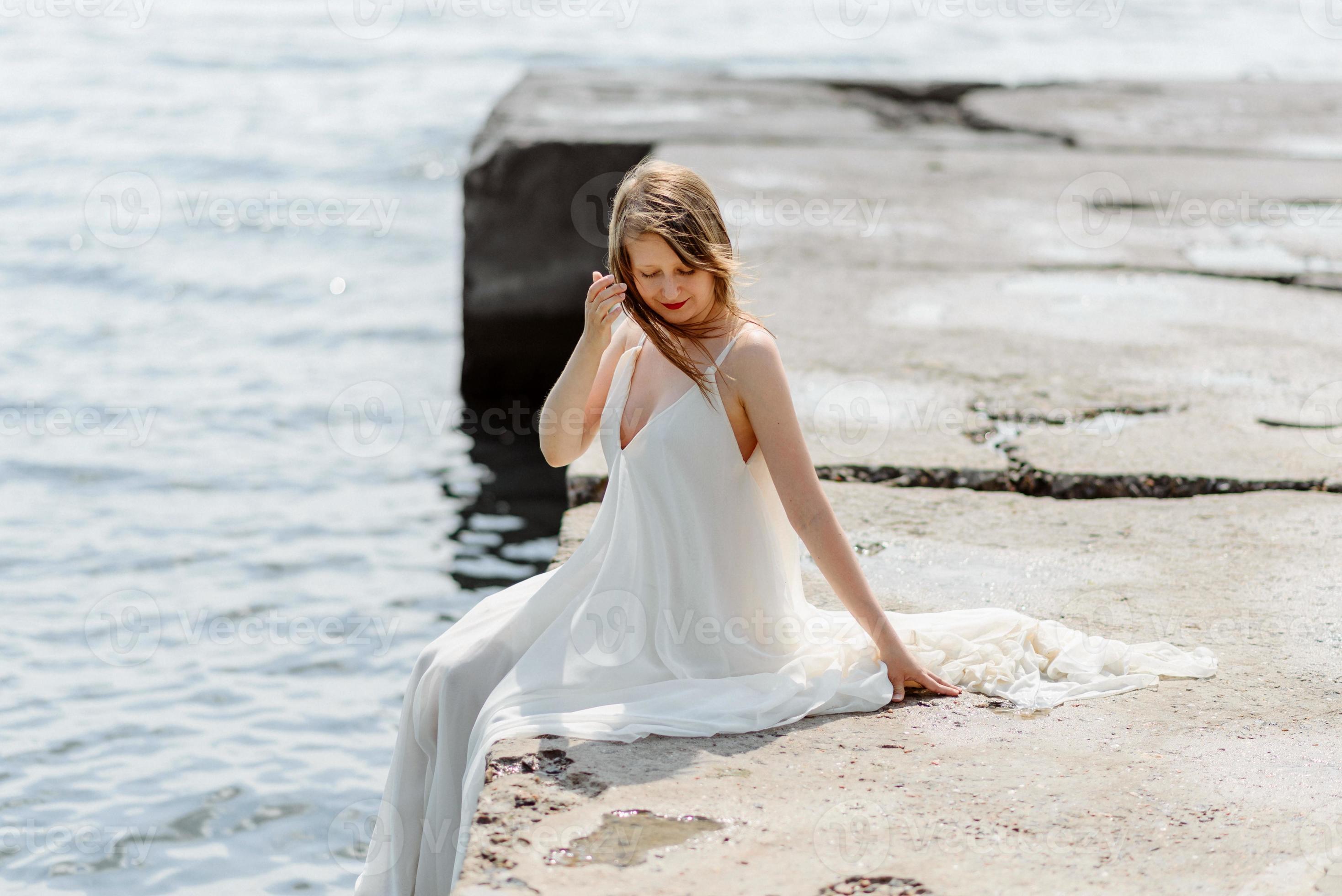 una giovane bella ragazza con un lungo vestito color latte cammina lungo la spiaggia e il molo sullo sfondo del mare. foto