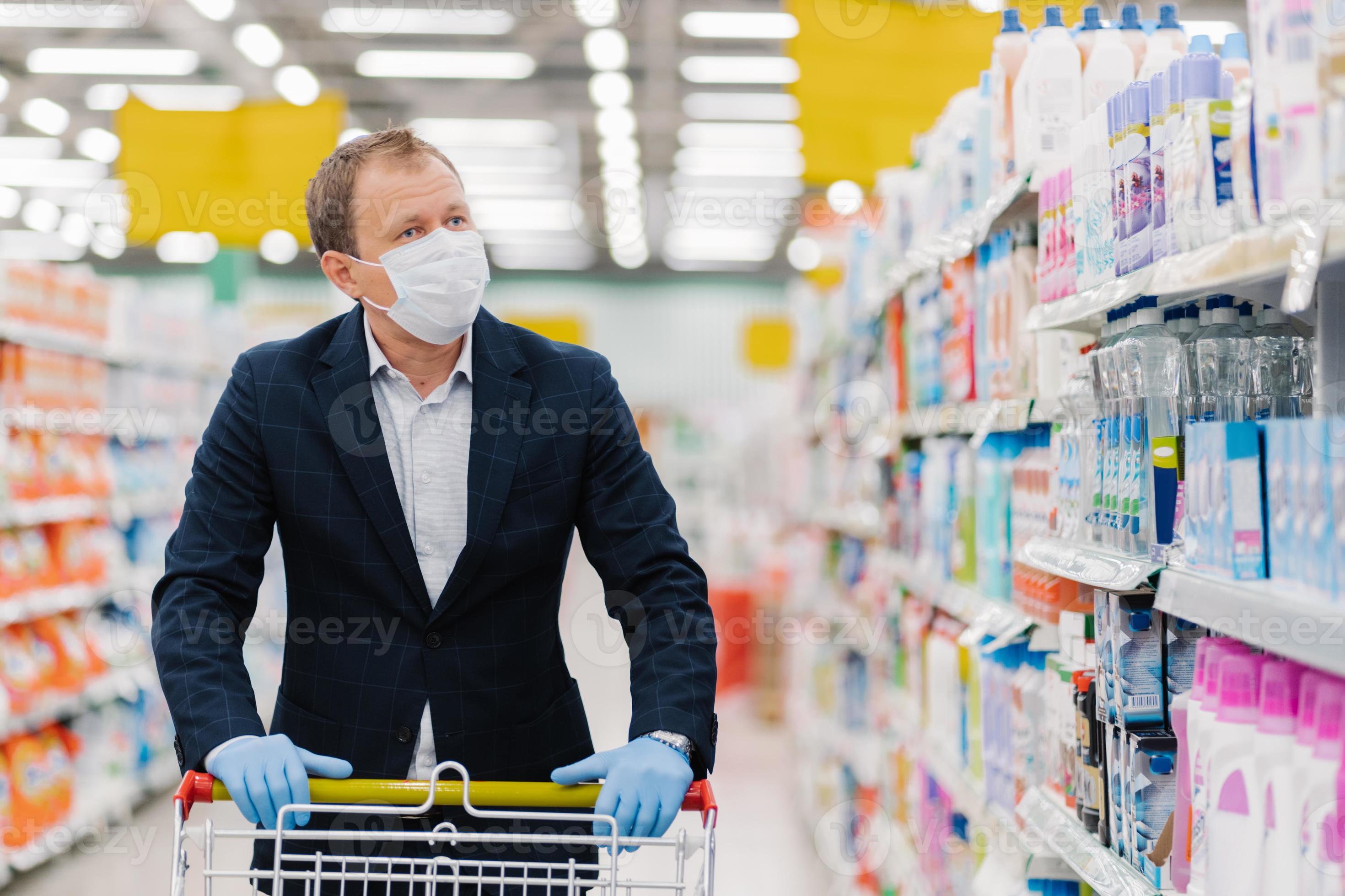 un uomo attento e serio sceglie i detersivi per la casa in negozio, cammina  con il carrello, acquista i prodotti necessari durante la quarantena,  indossa maschera medica protettiva e guanti. uomo d'affari