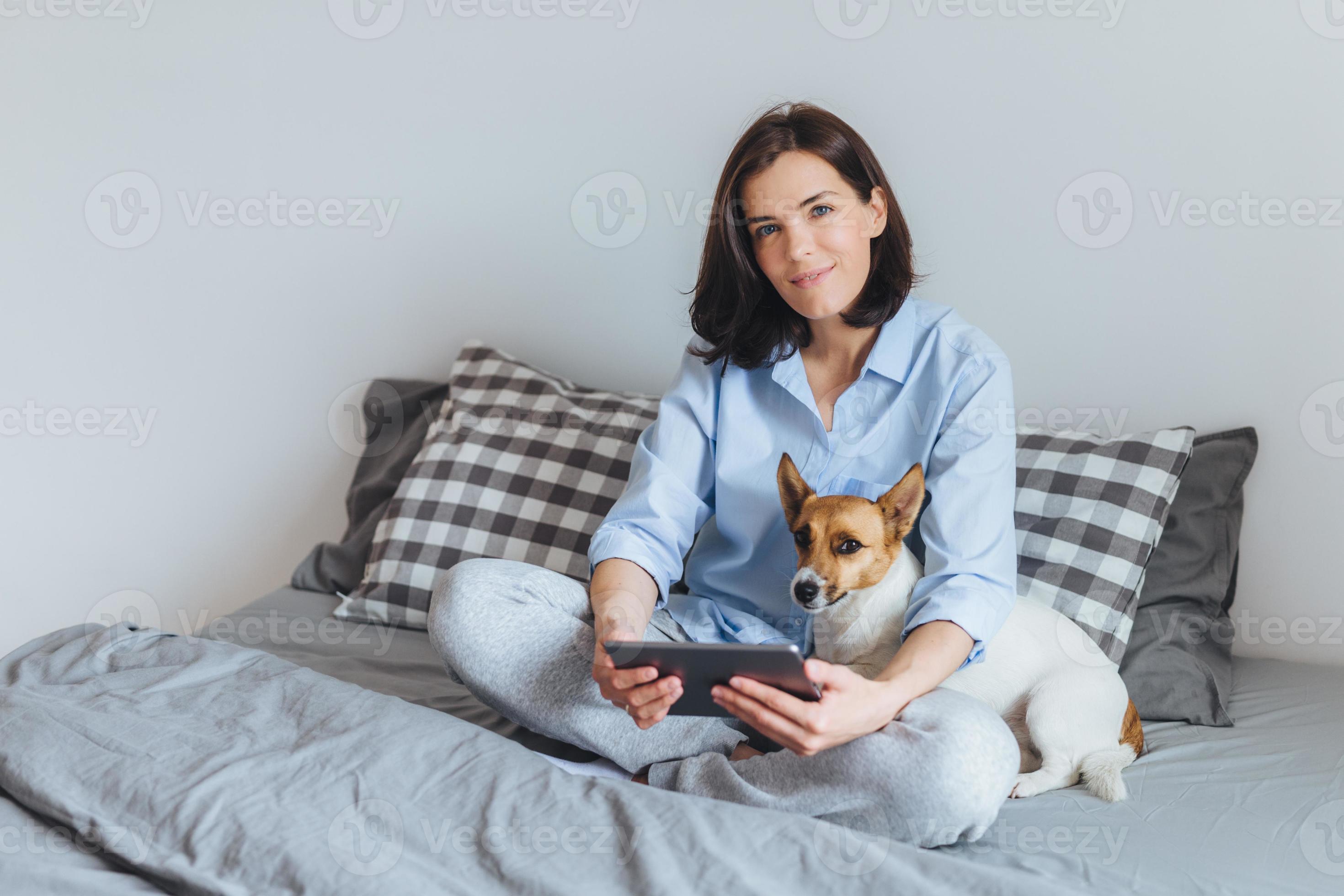 donna bruna dall'aspetto piacevole in pigiama usa gadget elettronici  moderni in camera da letto sul letto, si siede con il suo cane jack russell  terrier, gode di una calma atmosfera domestica, ha